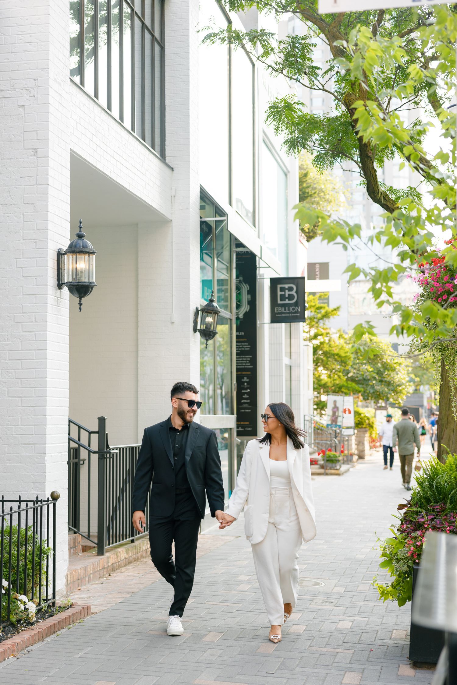 Stylish couple strolling hand in hand on a tree-lined sidewalk by white brick buildings.
