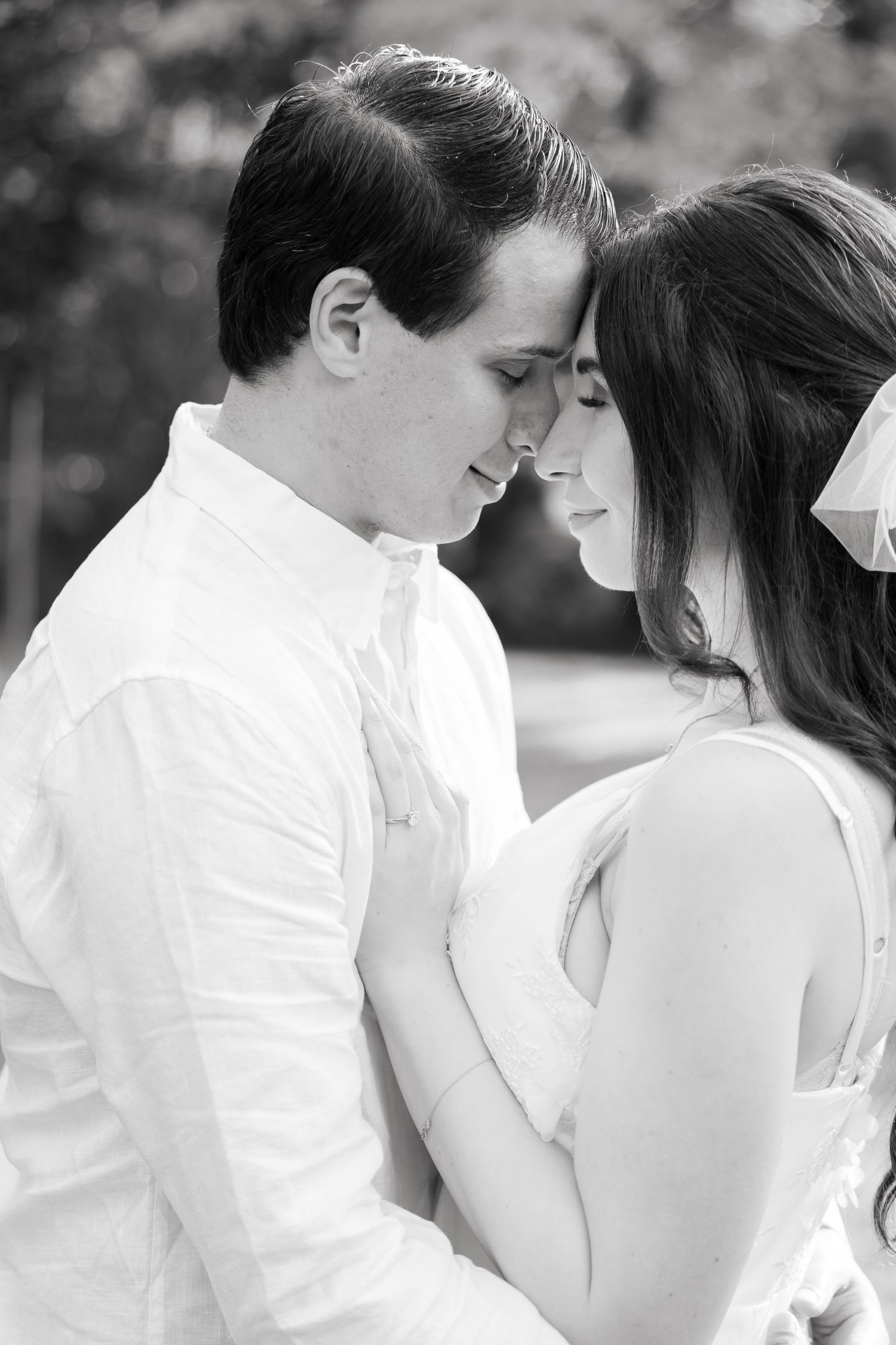 Intimate black and white portrait of a couple in wedding attire touching foreheads against a soft natural background.