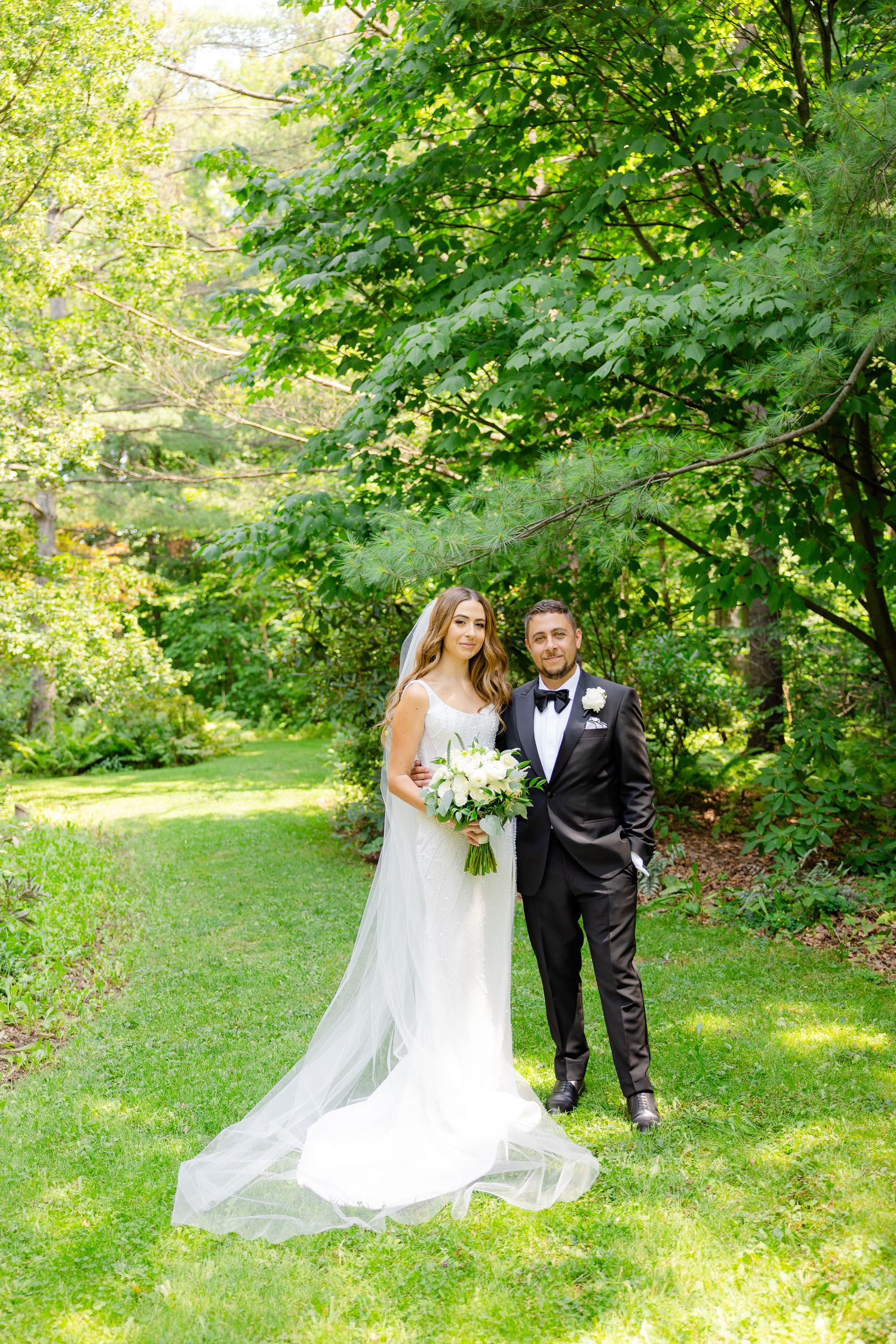 Wedding couple poses in lush green garden setting with bride holding white floral bouquet.