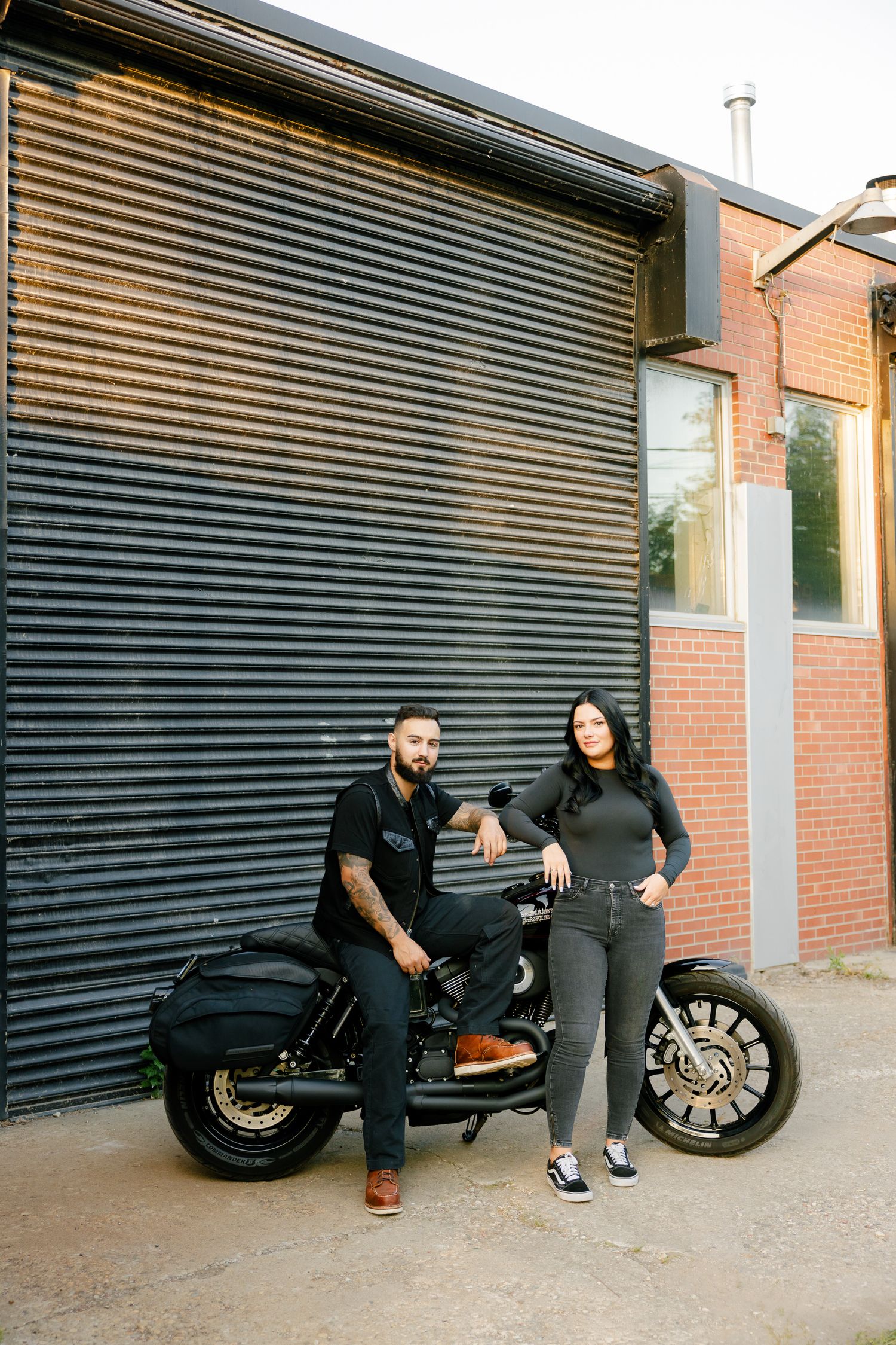 A couple poses with a custom motorcycle against an industrial metal garage door backdrop.