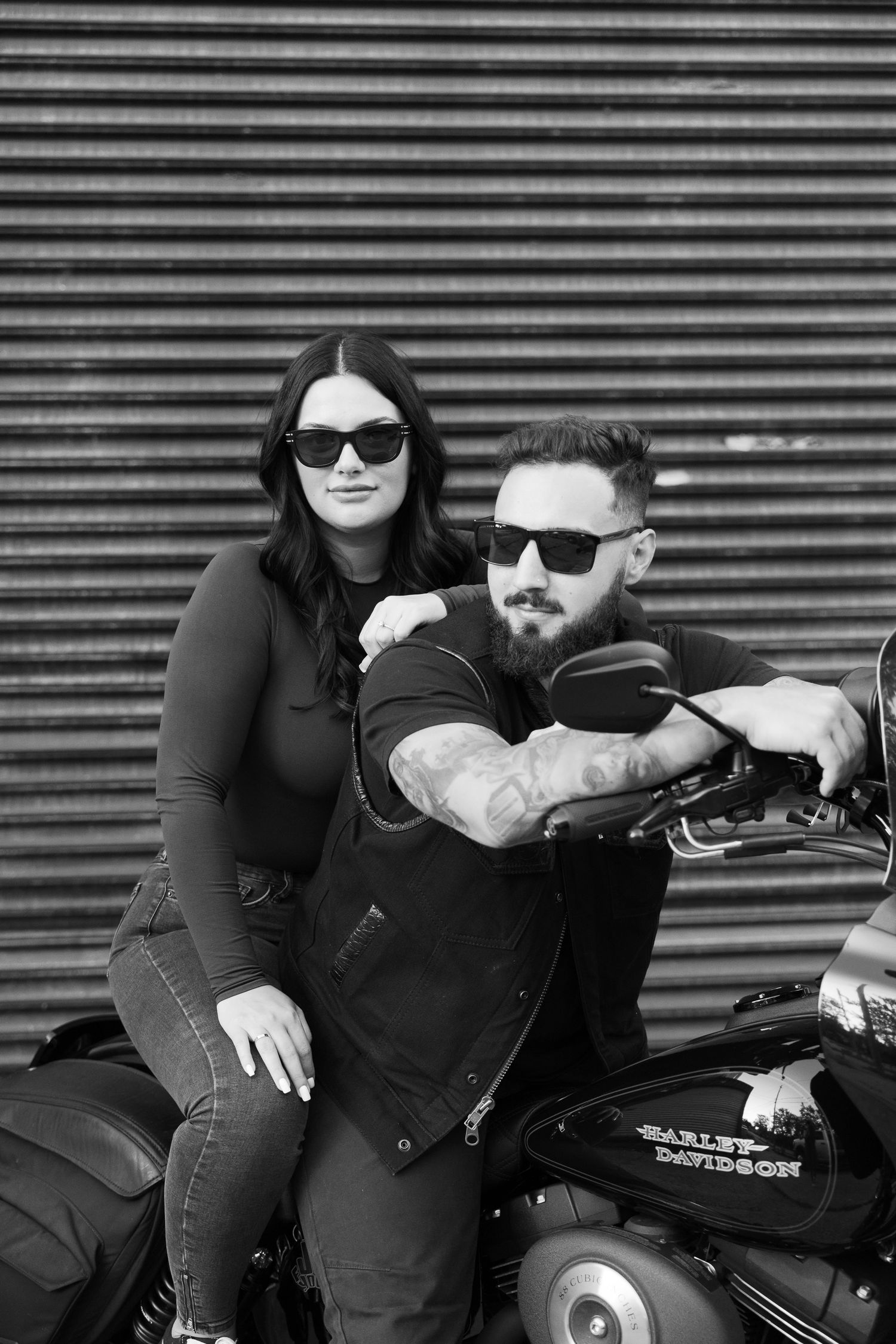 Black and white portrait of couple posing with motorcycle against metal wall.