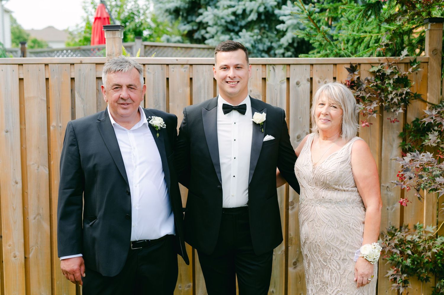 Three people in formal attire pose together at a backyard wedding celebration.