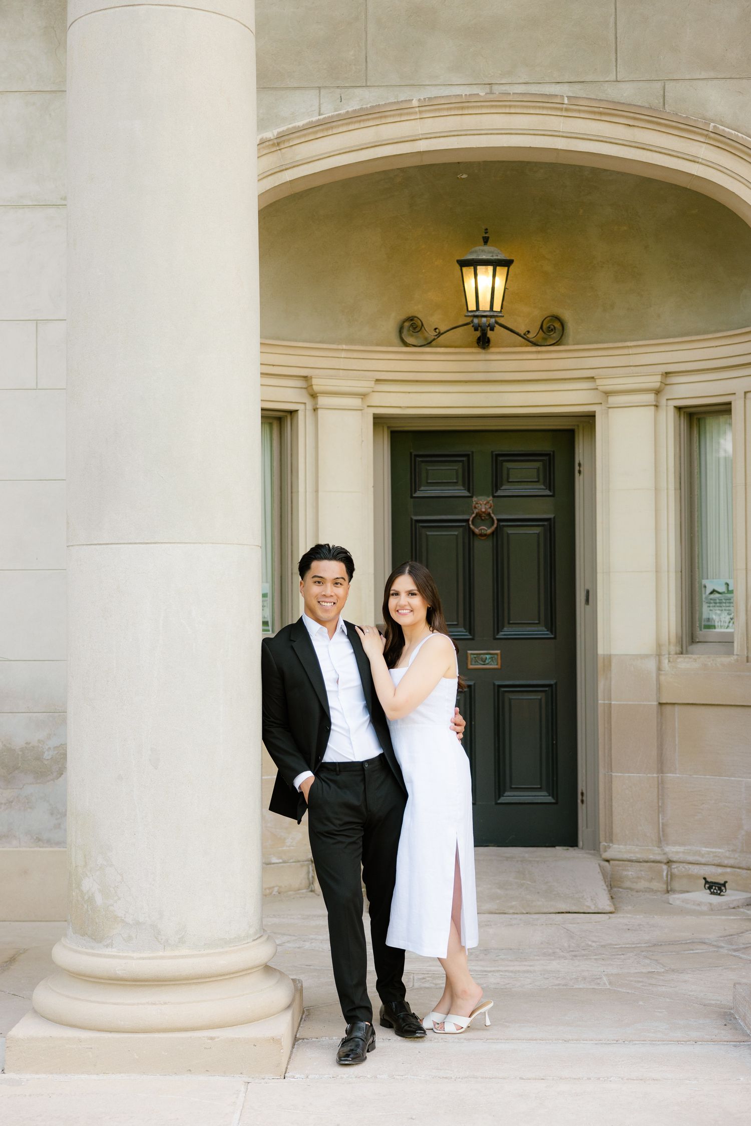 Elegant couple in formal attire pose together by historic building entrance with ornate lighting fixture.