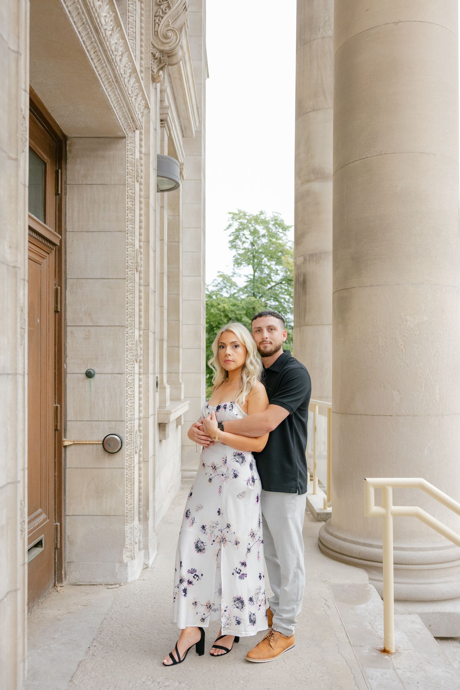 Couple in coordinated casual formal wear poses between grand stone columns.