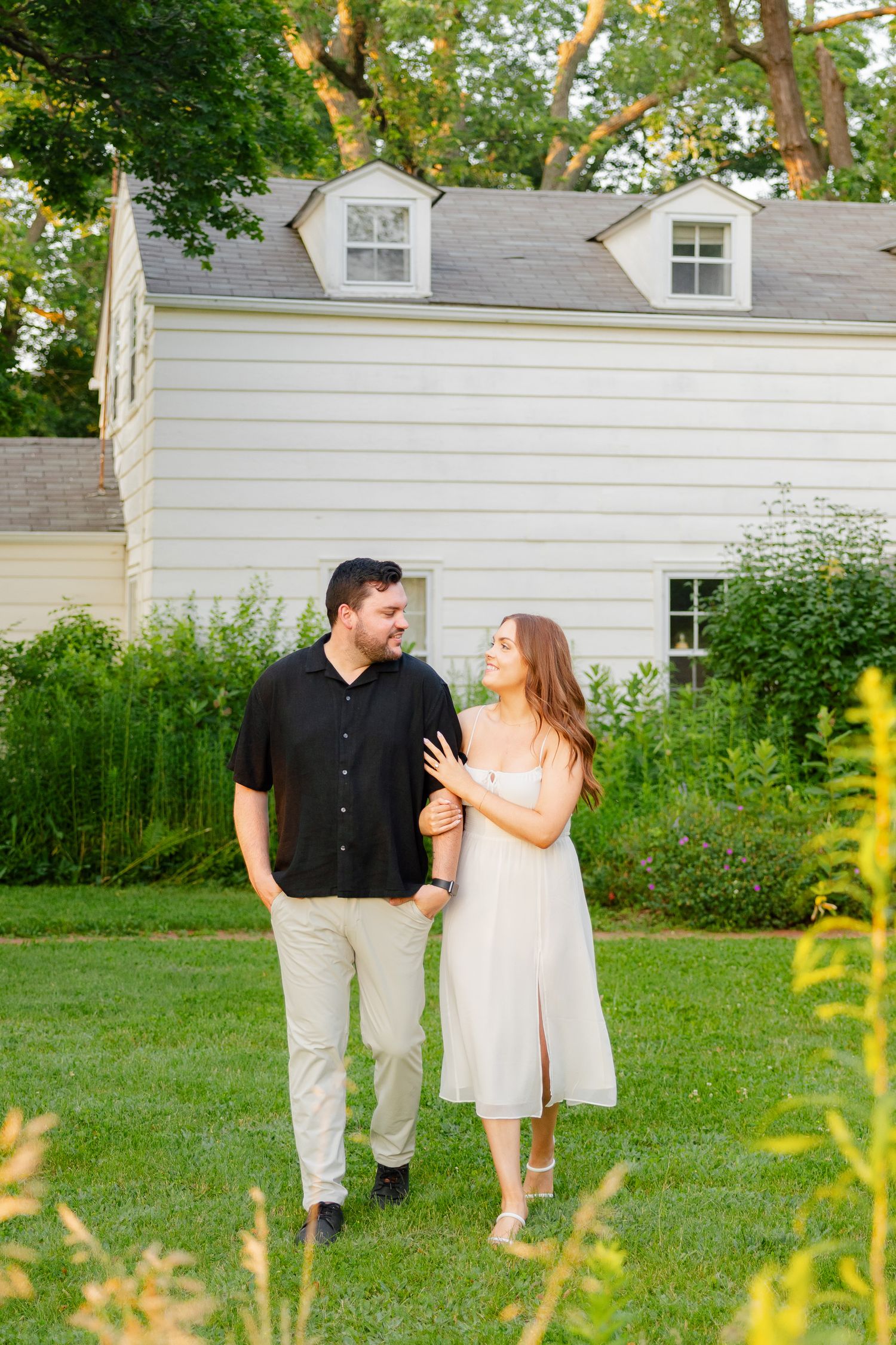Couple walking together on grass with white colonial house and summer foliage in background.