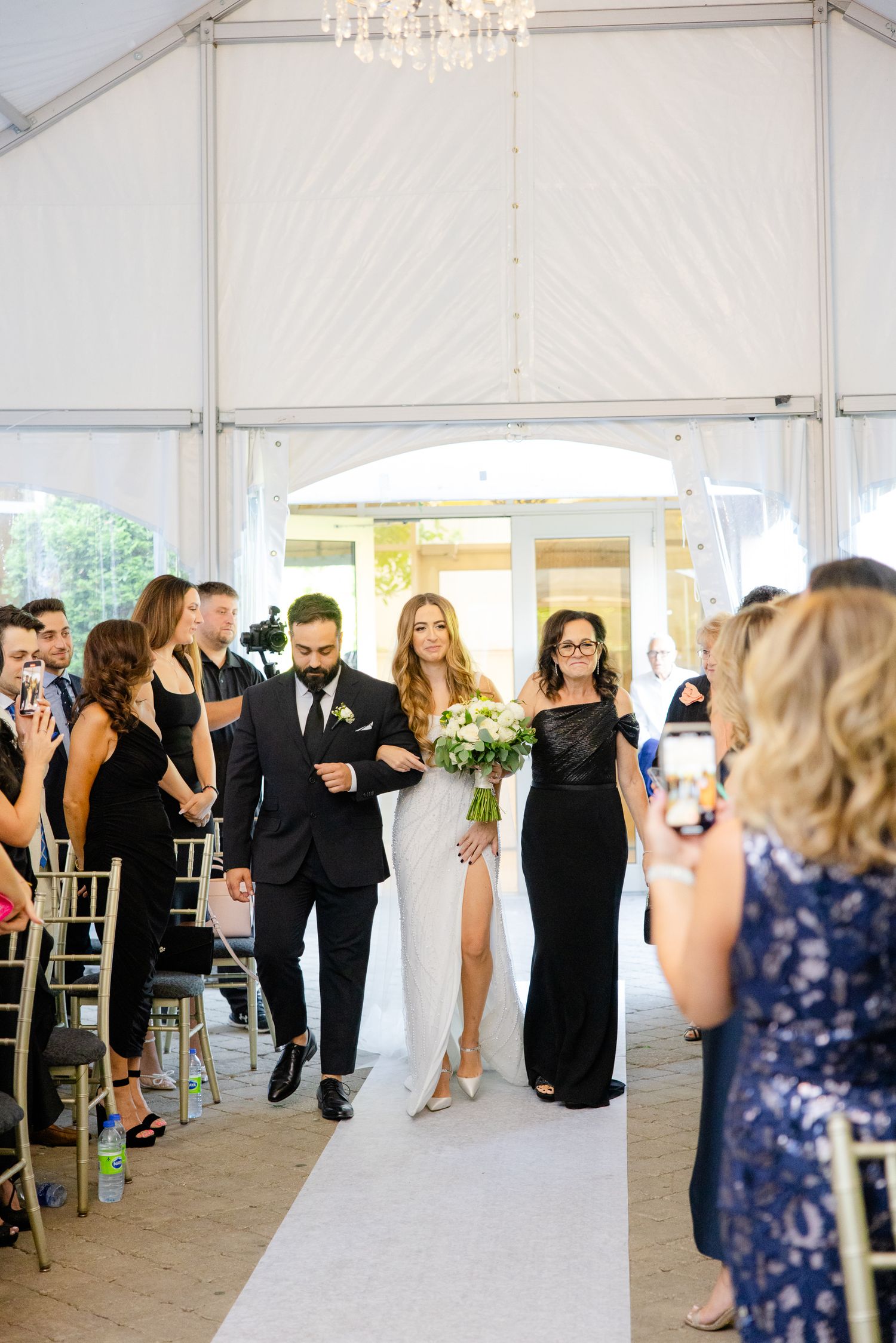 Wedding processional in an elegant white tent with guests seated along the aisle.