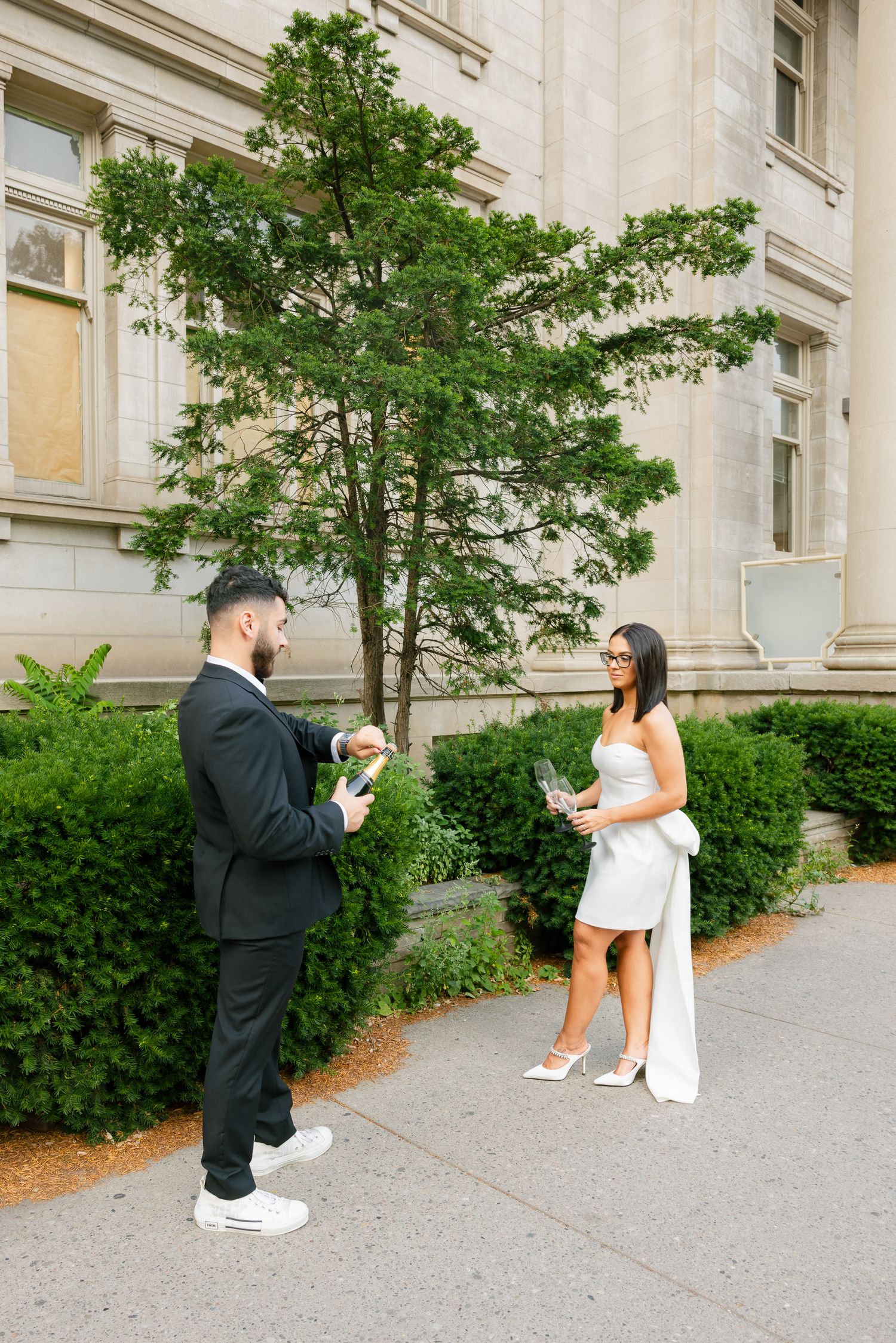 A couple stands in conversation outside an elegant building with landscaped greenery.