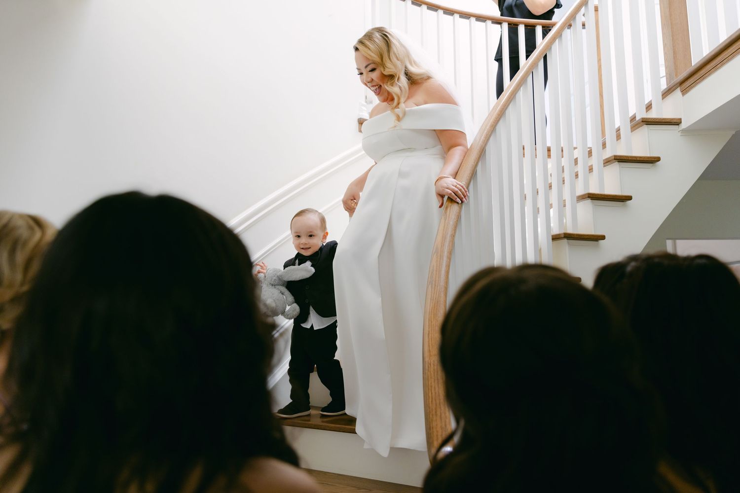 A bride descends a white staircase while holding hands with a small child in formal wear.