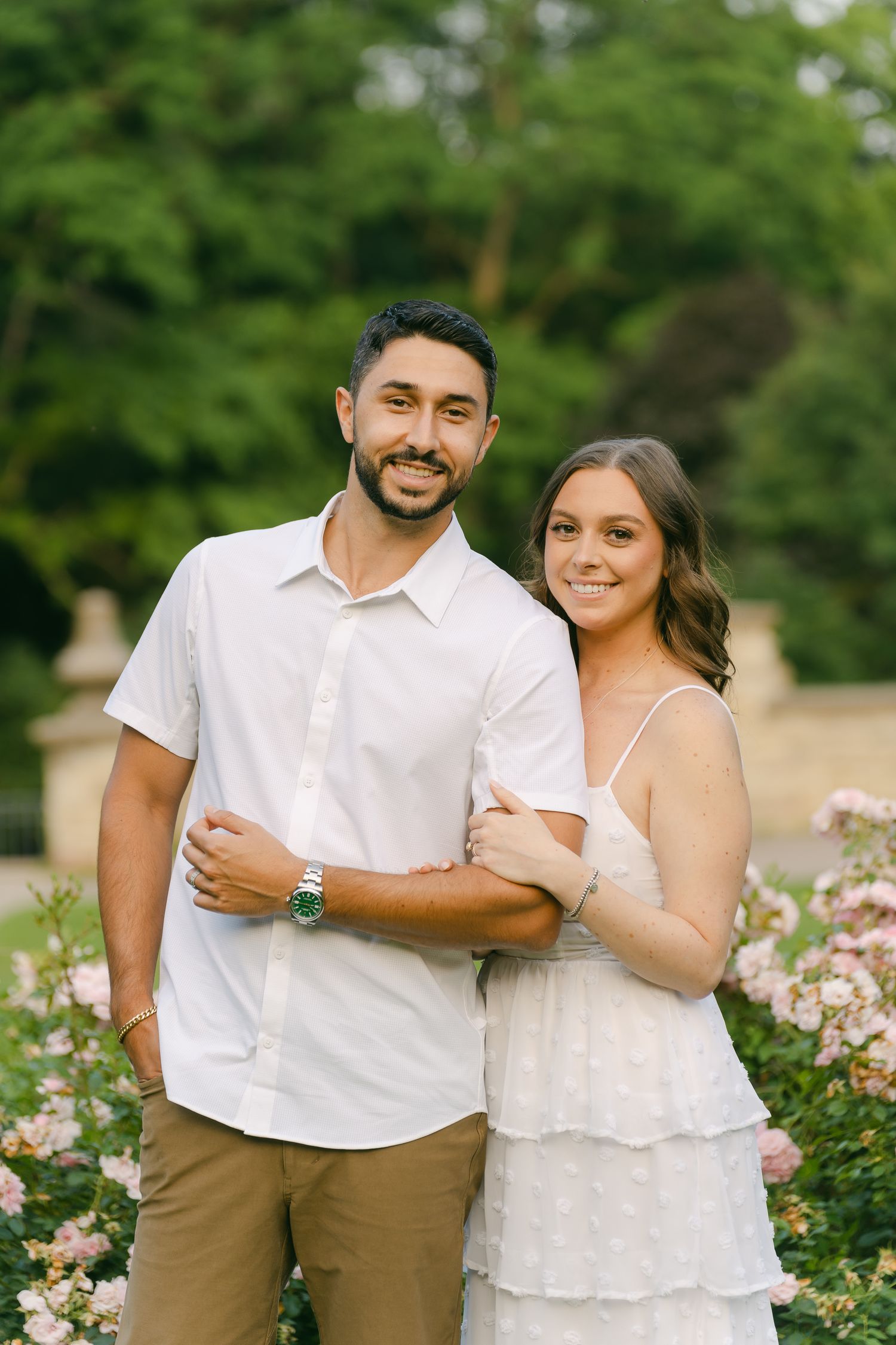 Happy couple in coordinated white outfits pose in front of flowering rose bushes.