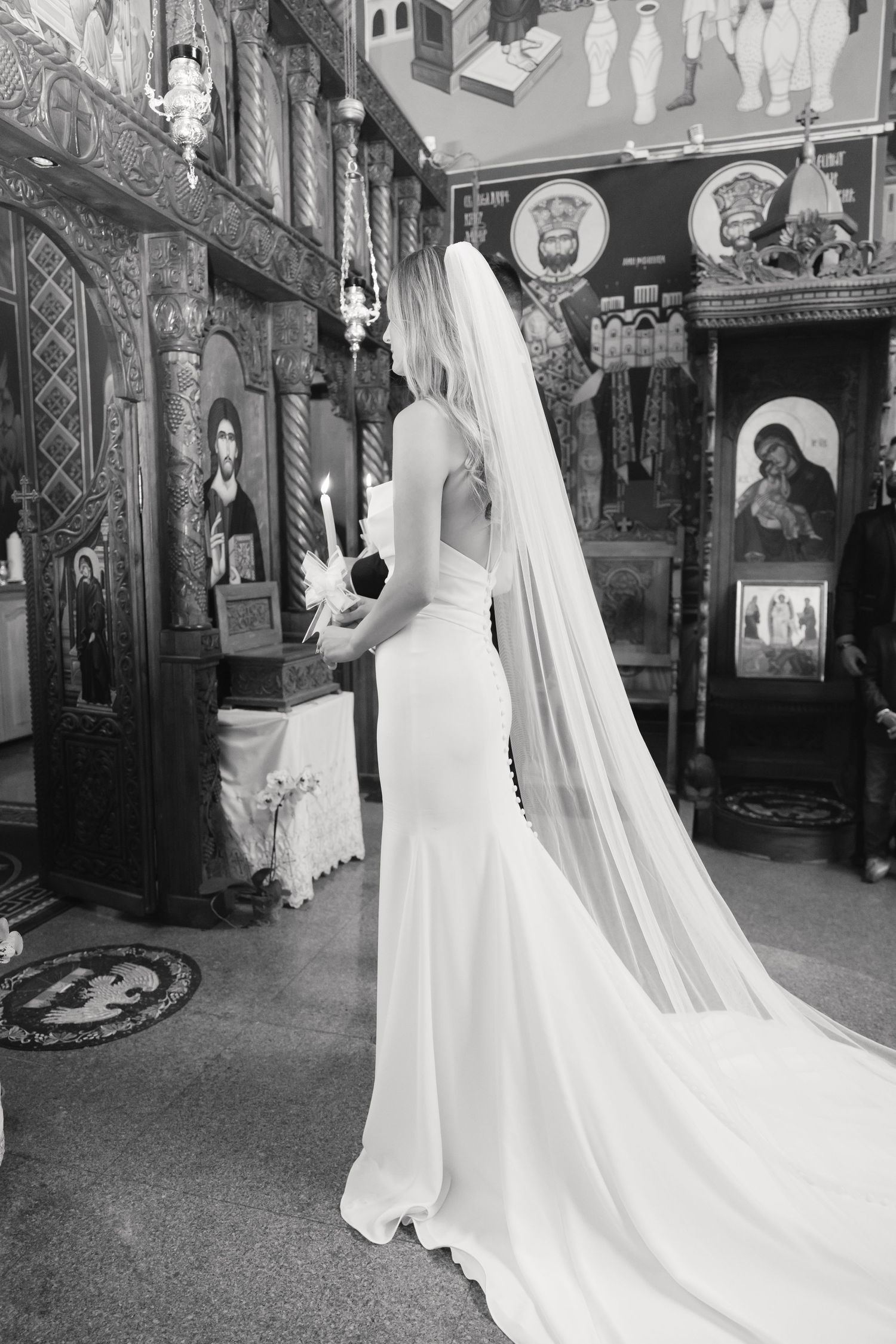 Elegant bride in form-fitting white gown with flowing veil stands in ornately decorated Orthodox church.