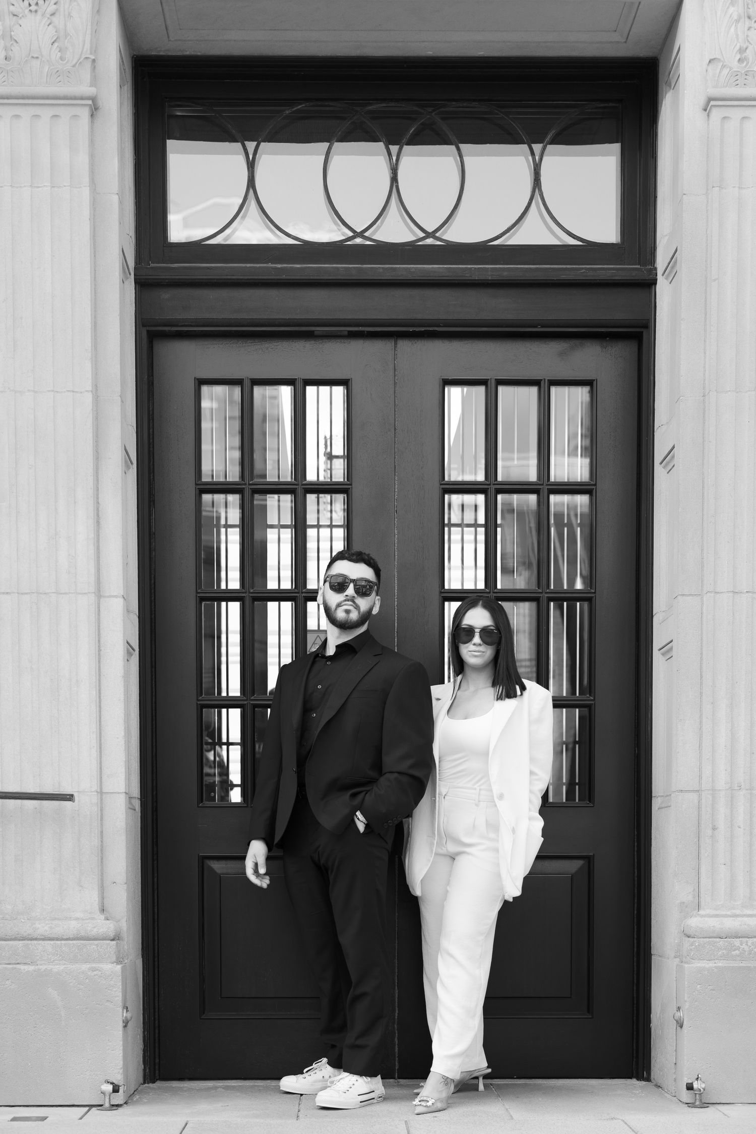 Stylishly dressed couple in black and white outfits pose in front of elegant double doors in monochrome photo.