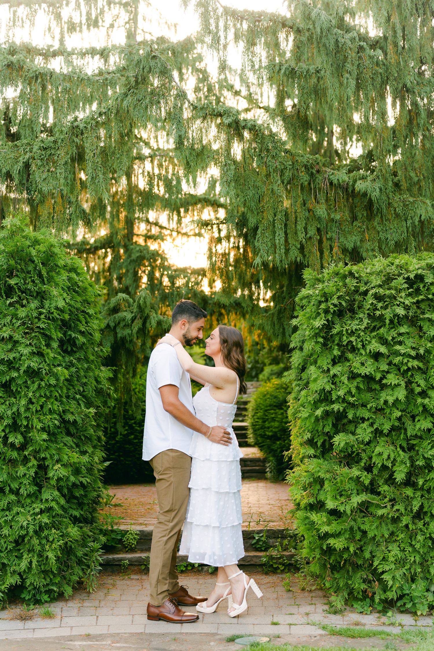 Couple in summer attire poses romantically on garden steps between perfectly trimmed hedges.