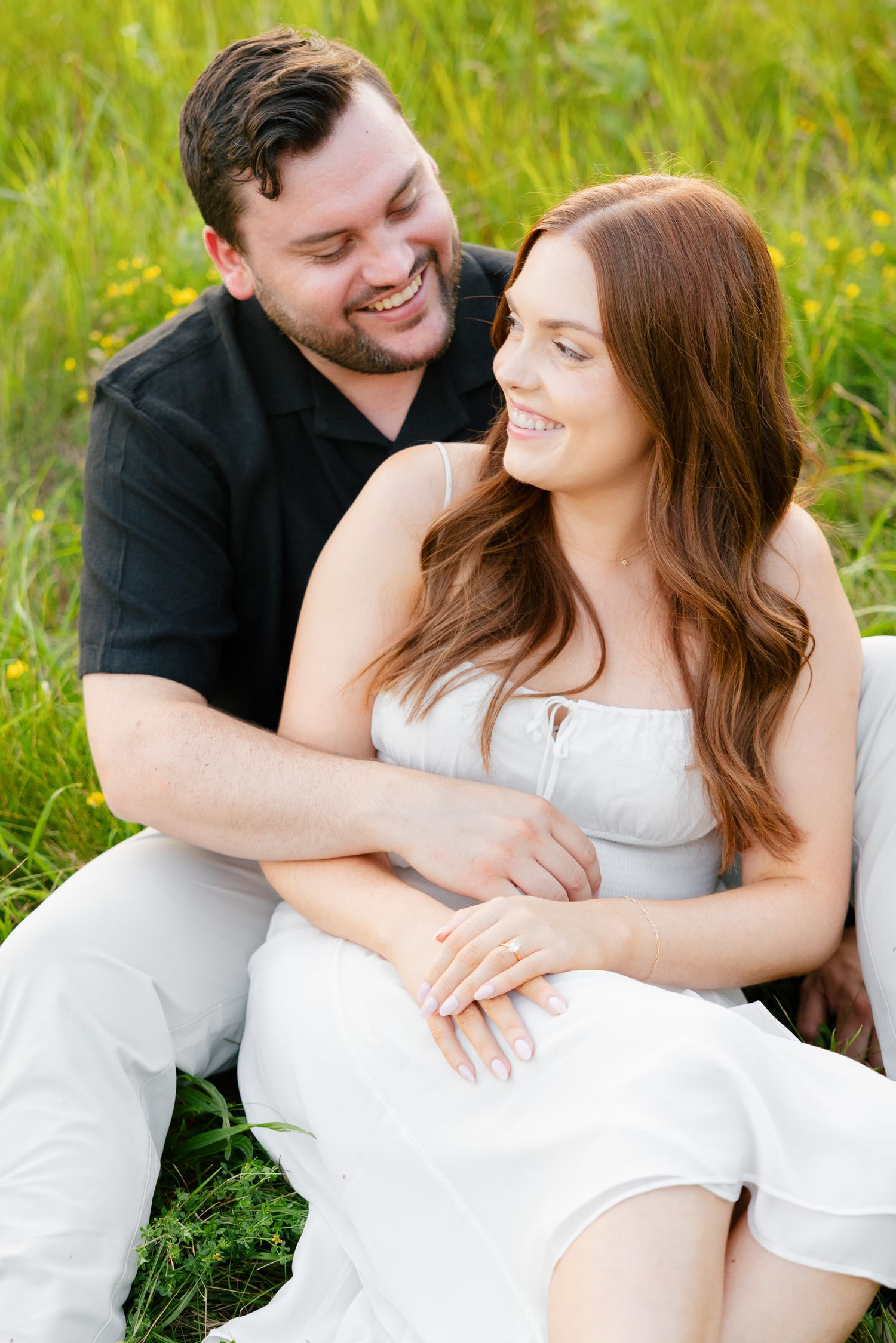 Joyful couple embraces while sitting in tall grass during an outdoor photo session.