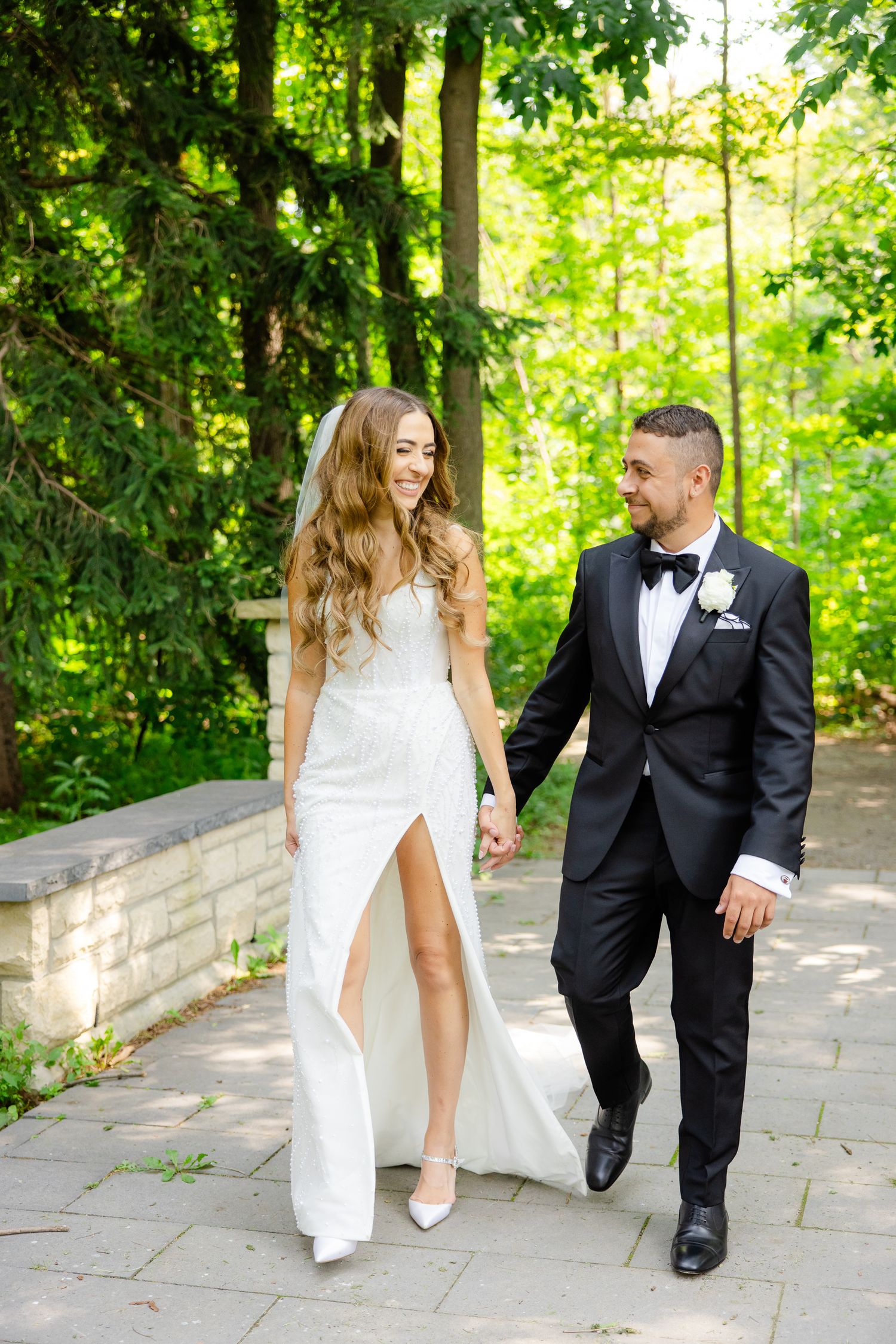 Elegant couple walking hand in hand along garden path with lush greenery backdrop.