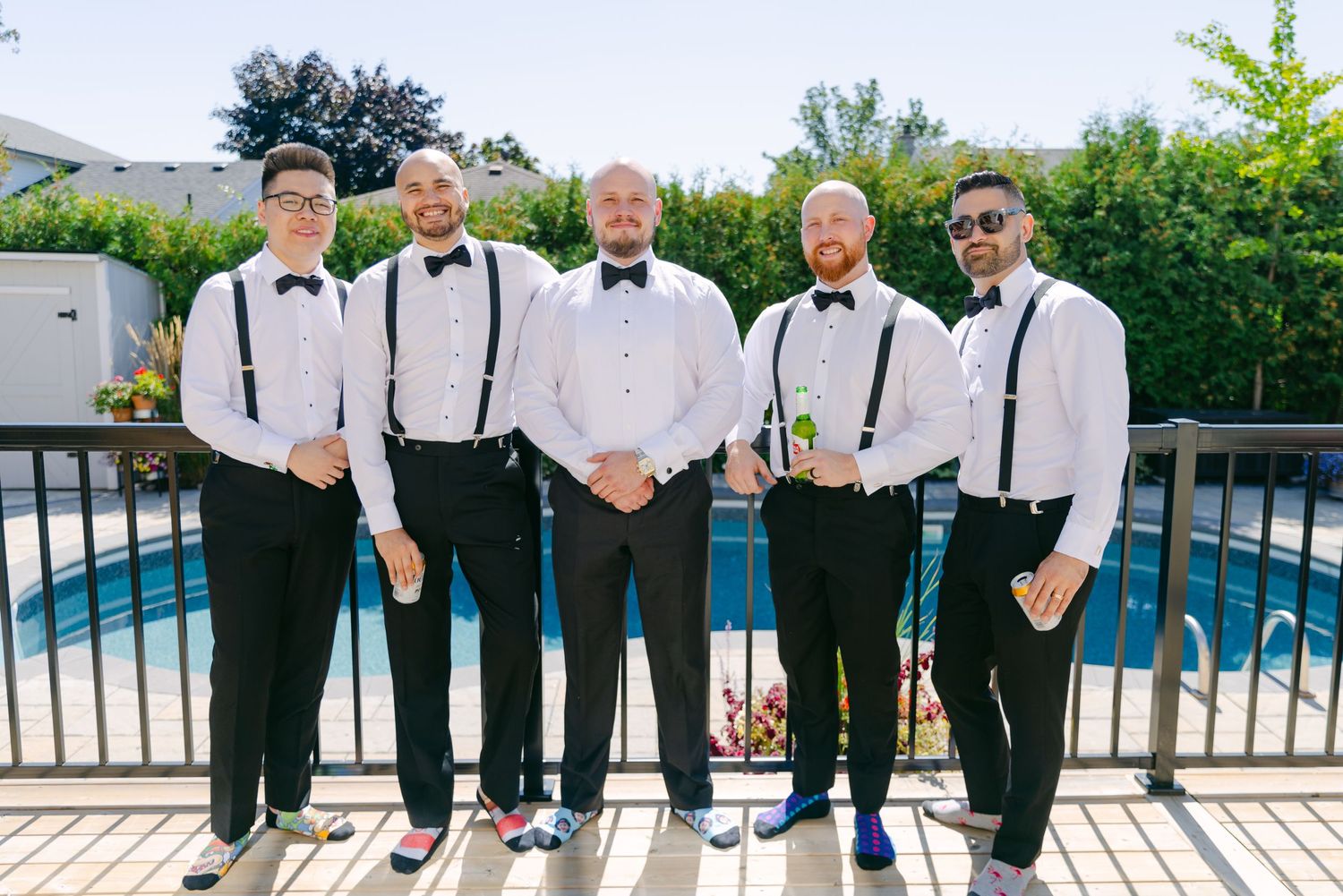 Group photo of wedding party in formal attire standing by pool with black bow ties and suspenders.