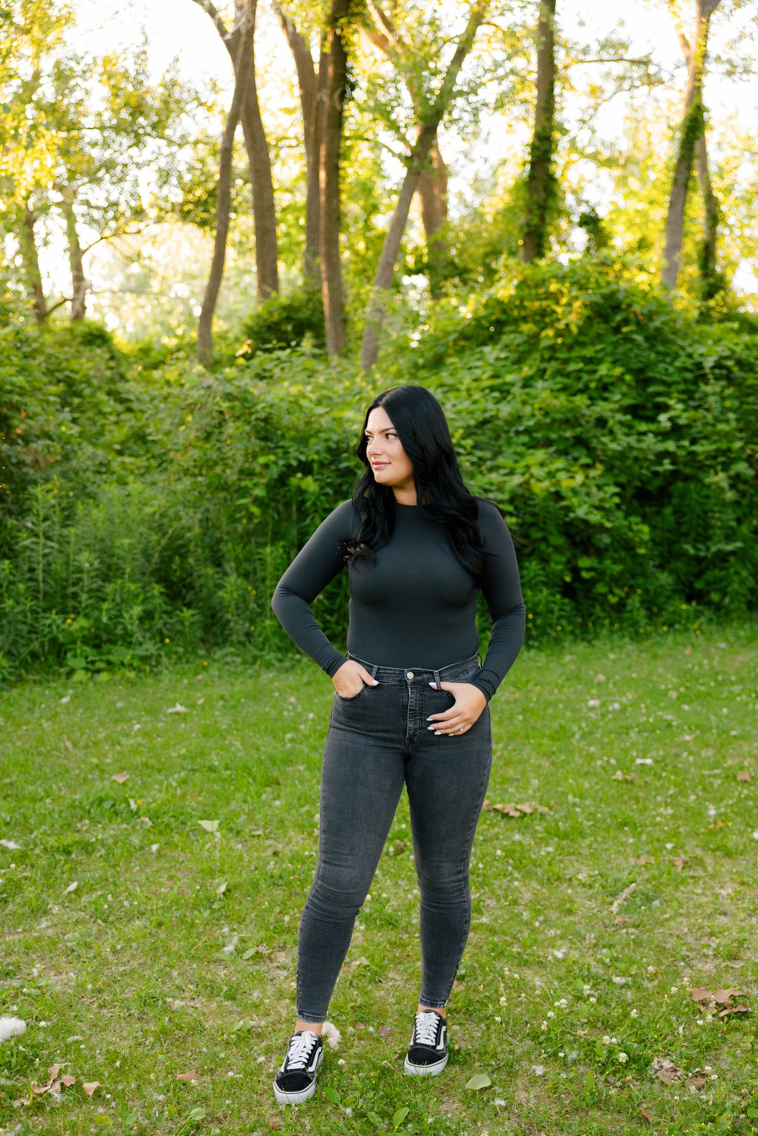 Woman in casual black outfit poses naturally in sunny woodland clearing.