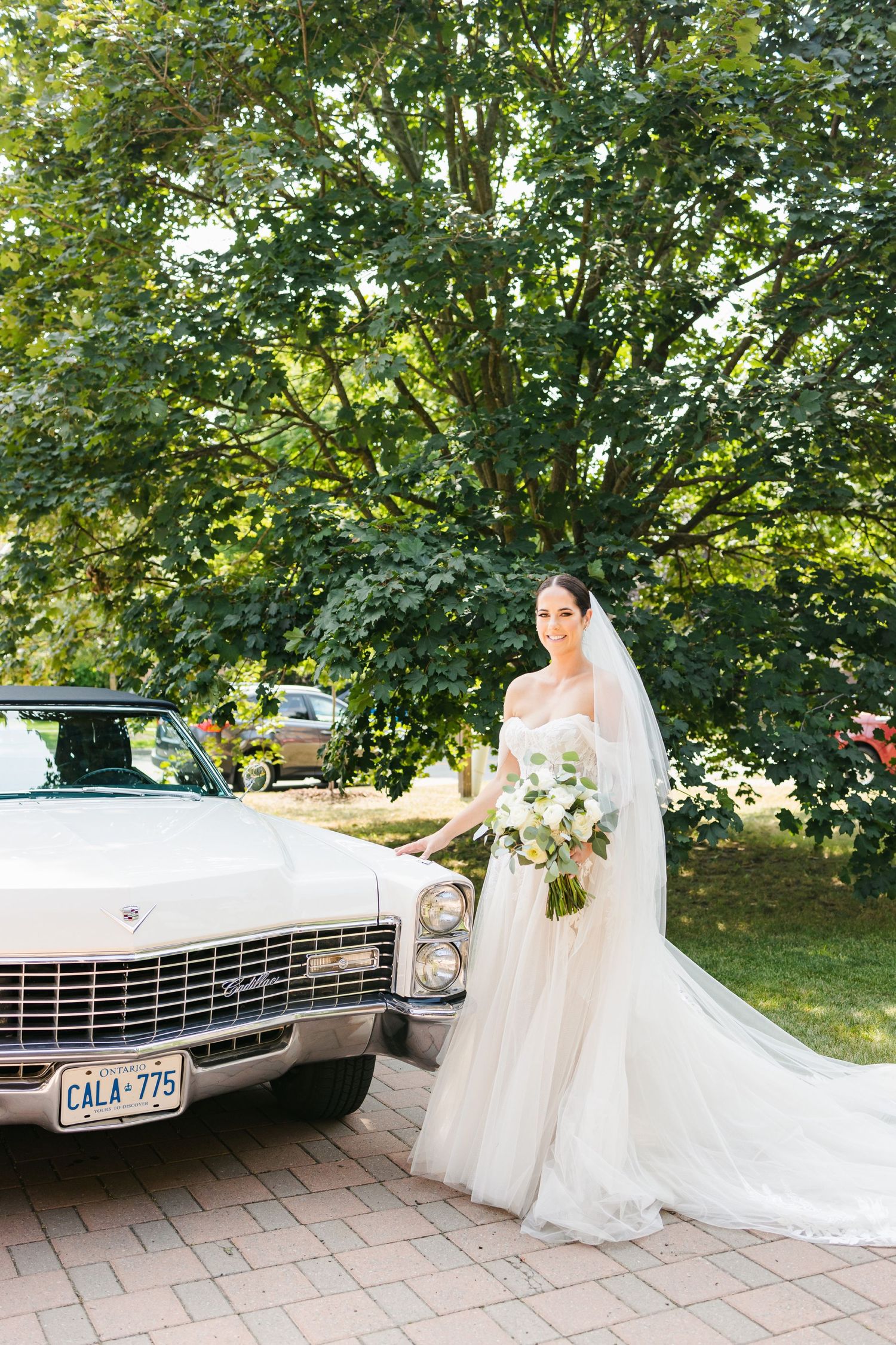 Beautiful bride in flowing gown poses beside vintage white Cadillac in garden setting.