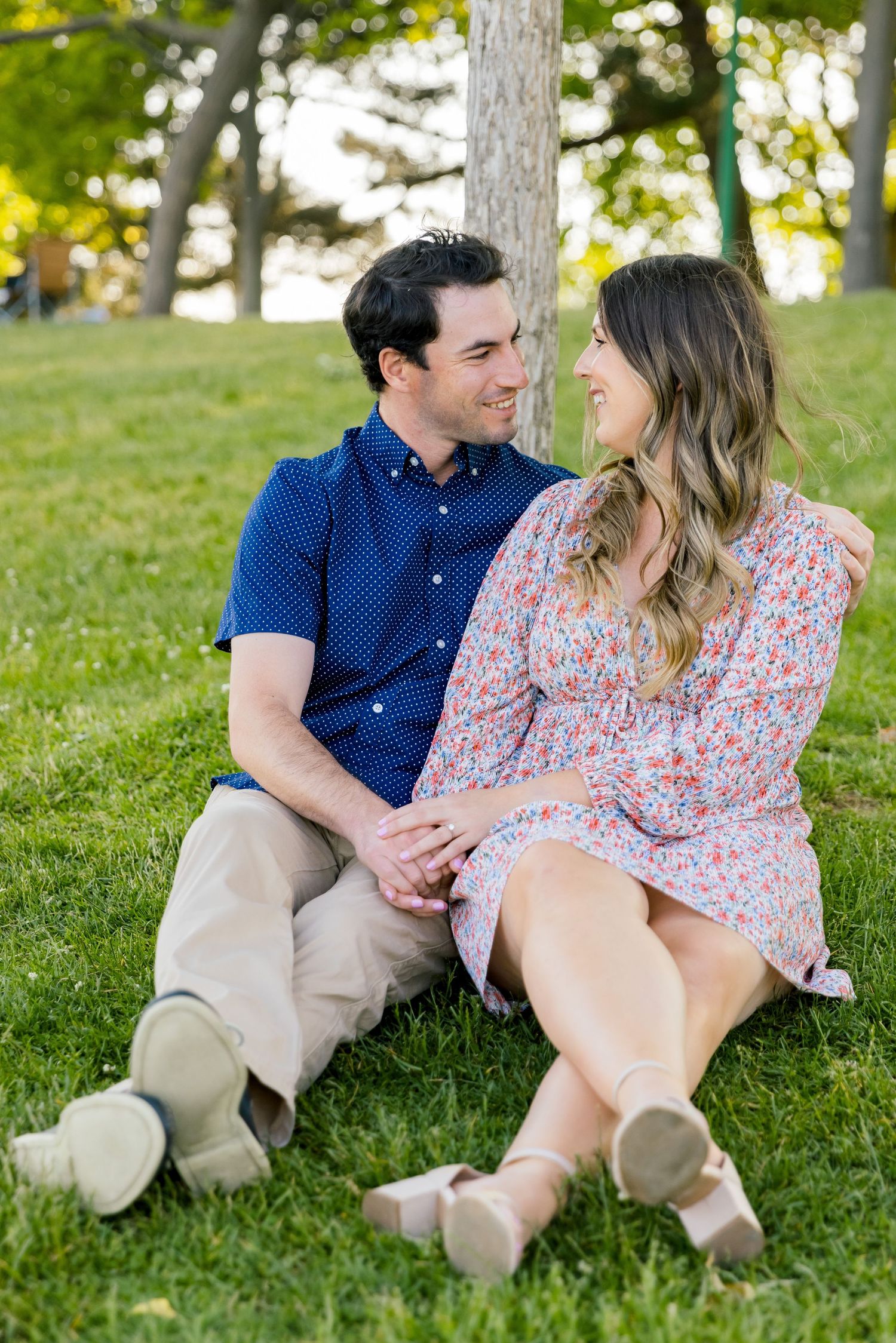 A couple shares a tender moment while sitting on grass beneath a tree.