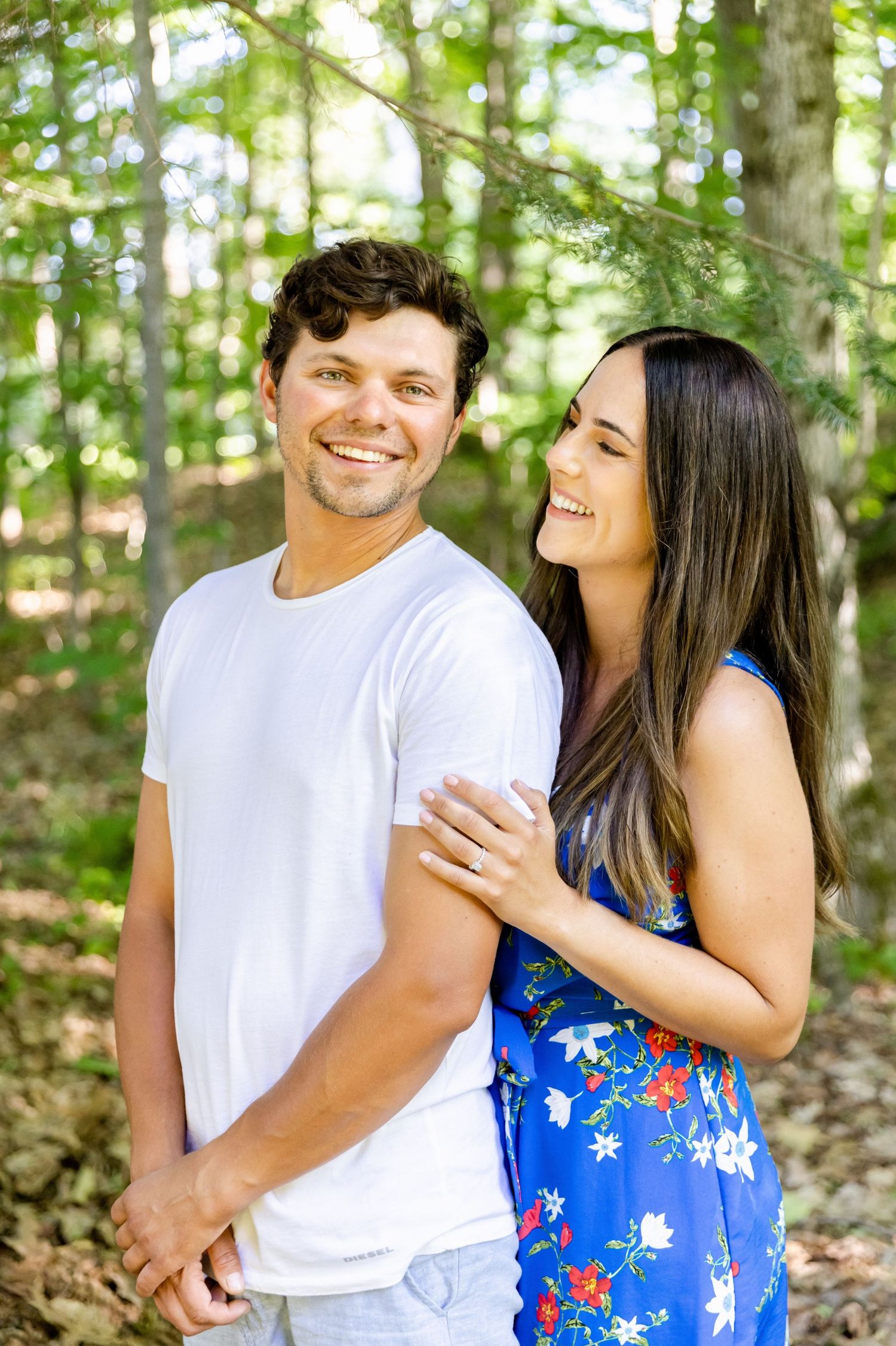 A couple embraces in a sunlit forest setting, with her wearing a blue floral dress and him in a white t-shirt.