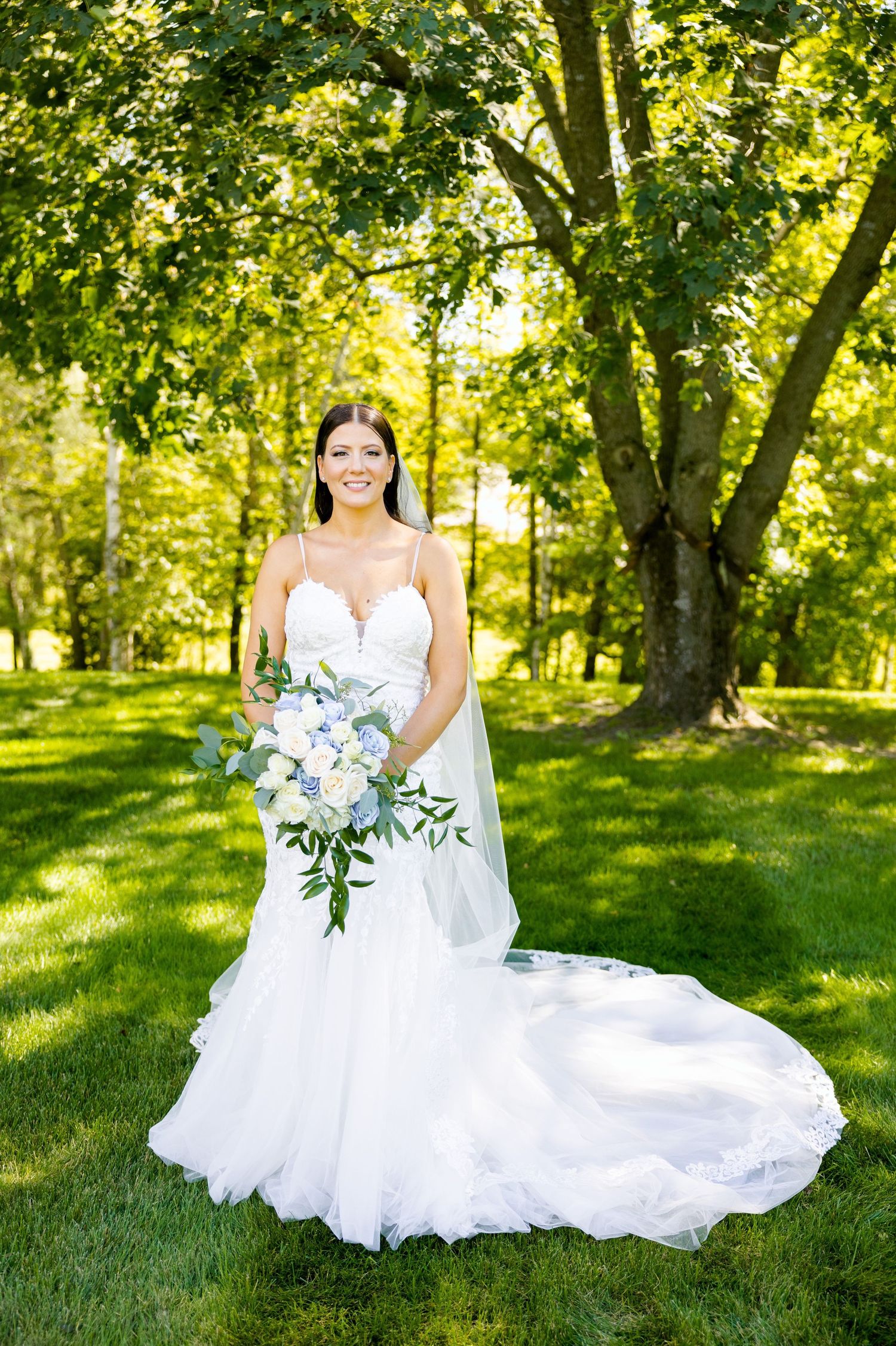 A bride in a flowing white dress holds a blue and white bouquet in a sunlit garden setting.