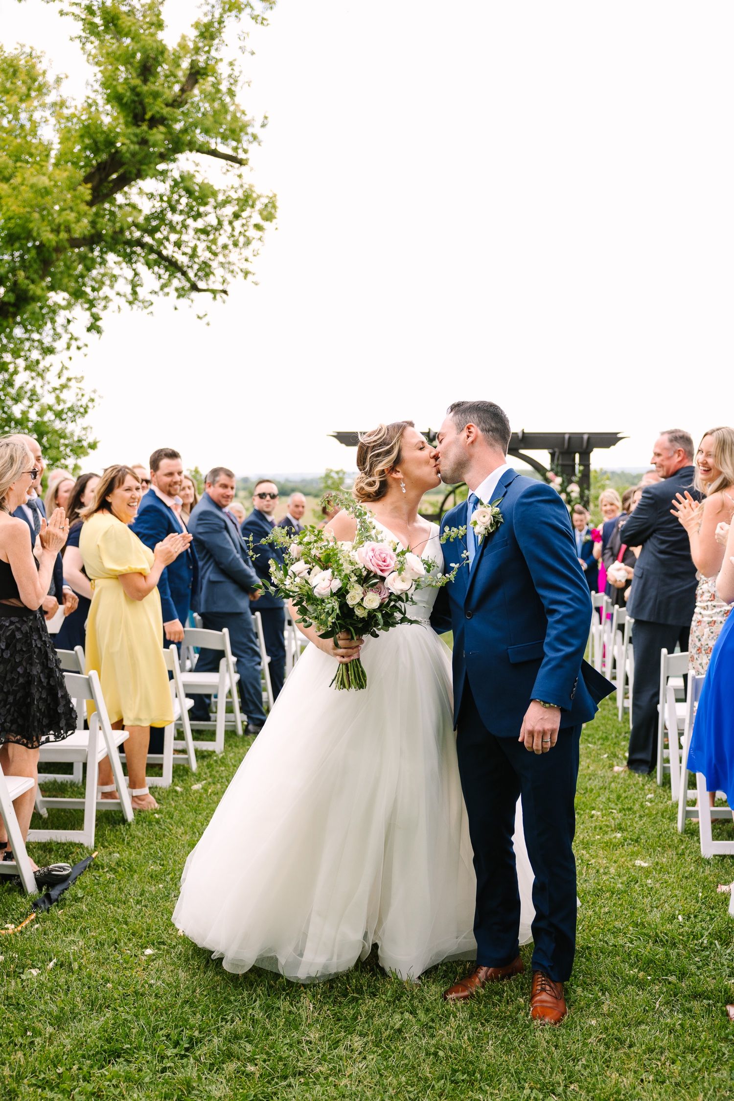 Wedding couple shares first kiss as guests cheer during outdoor ceremony with white chairs.