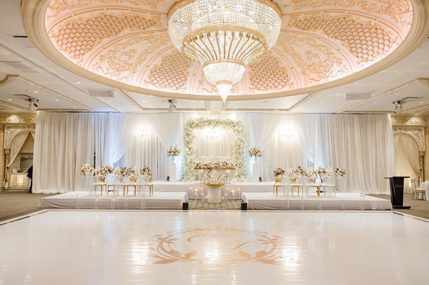 Luxurious white and gold ballroom with ornate ceiling, crystal chandelier and elegant wedding setup.