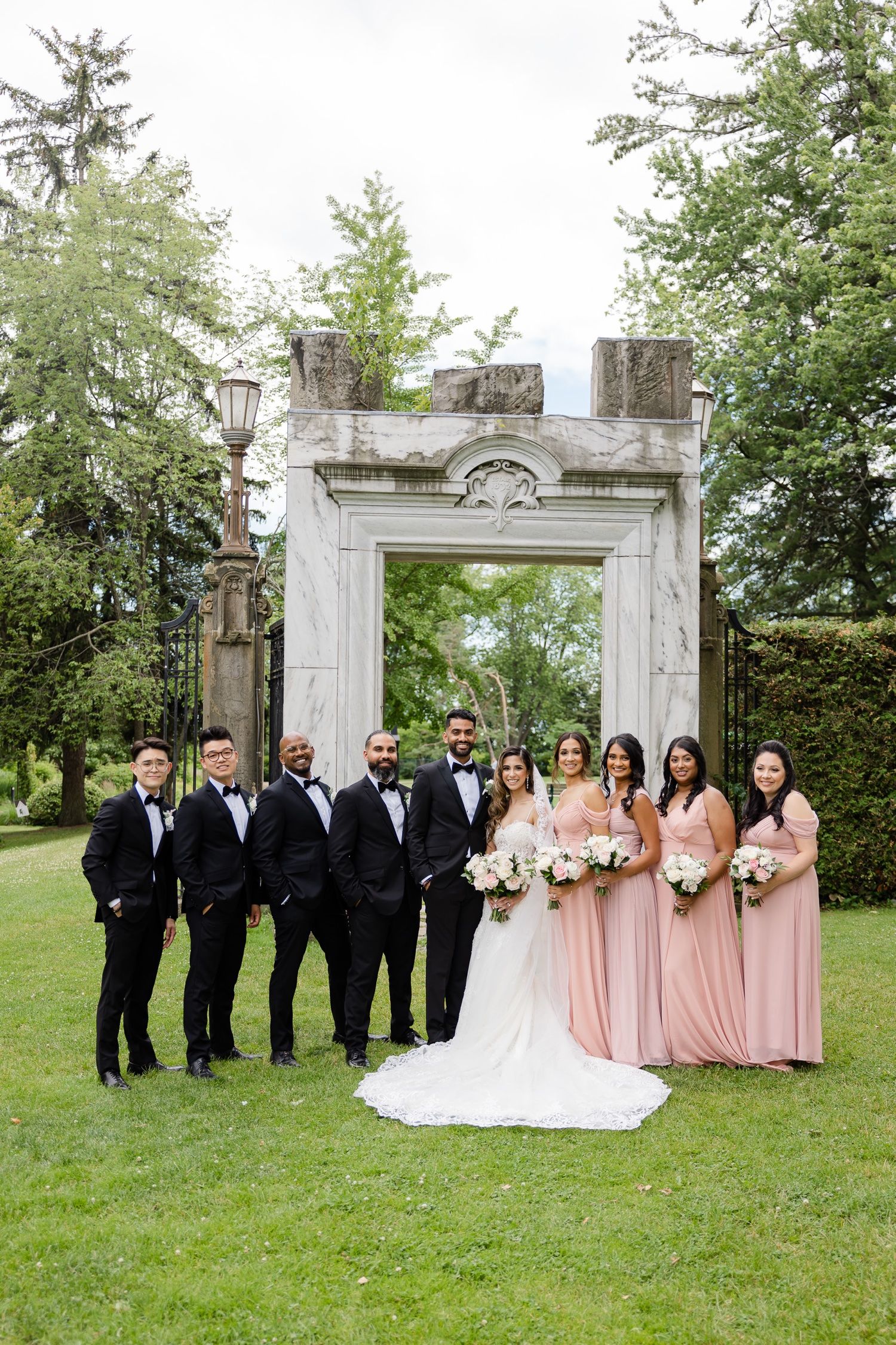 Formal wedding party portrait under a grand stone archway with bridesmaids in blush gowns and groomsmen in black suits.