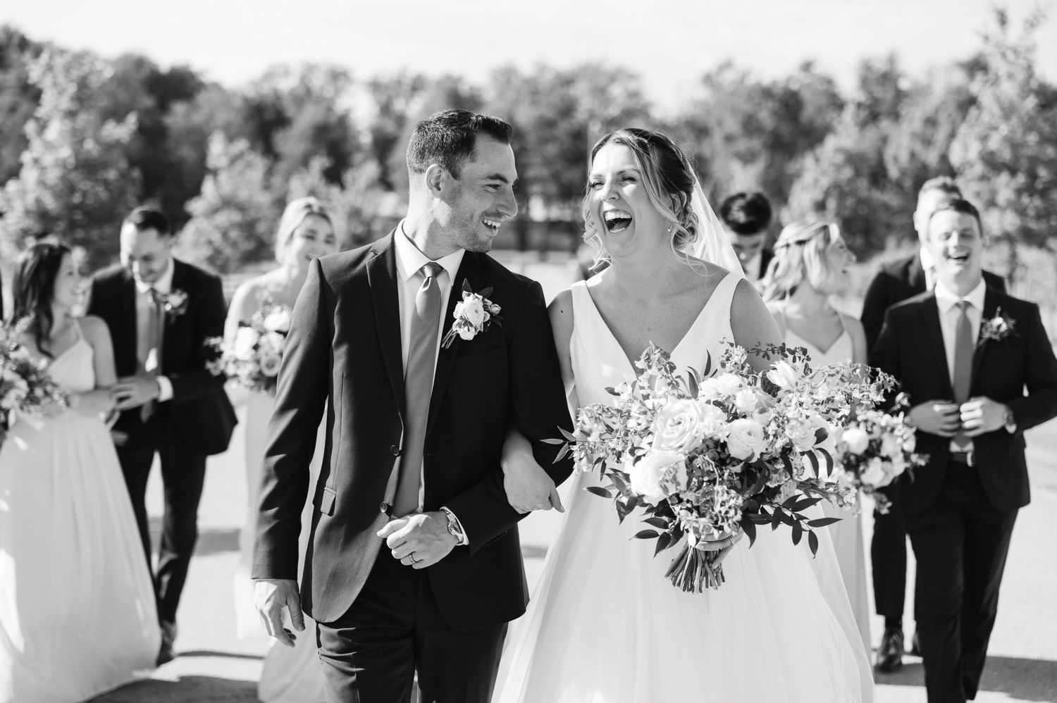 Black and white wedding photo showing joyful couple and wedding party on outdoor path.