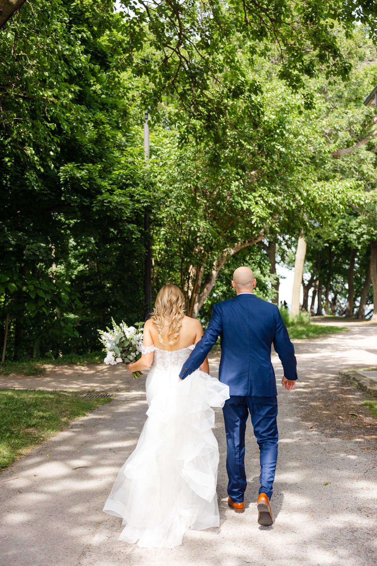 Newlyweds walking hand in hand down tree-lined path in formal attire.
