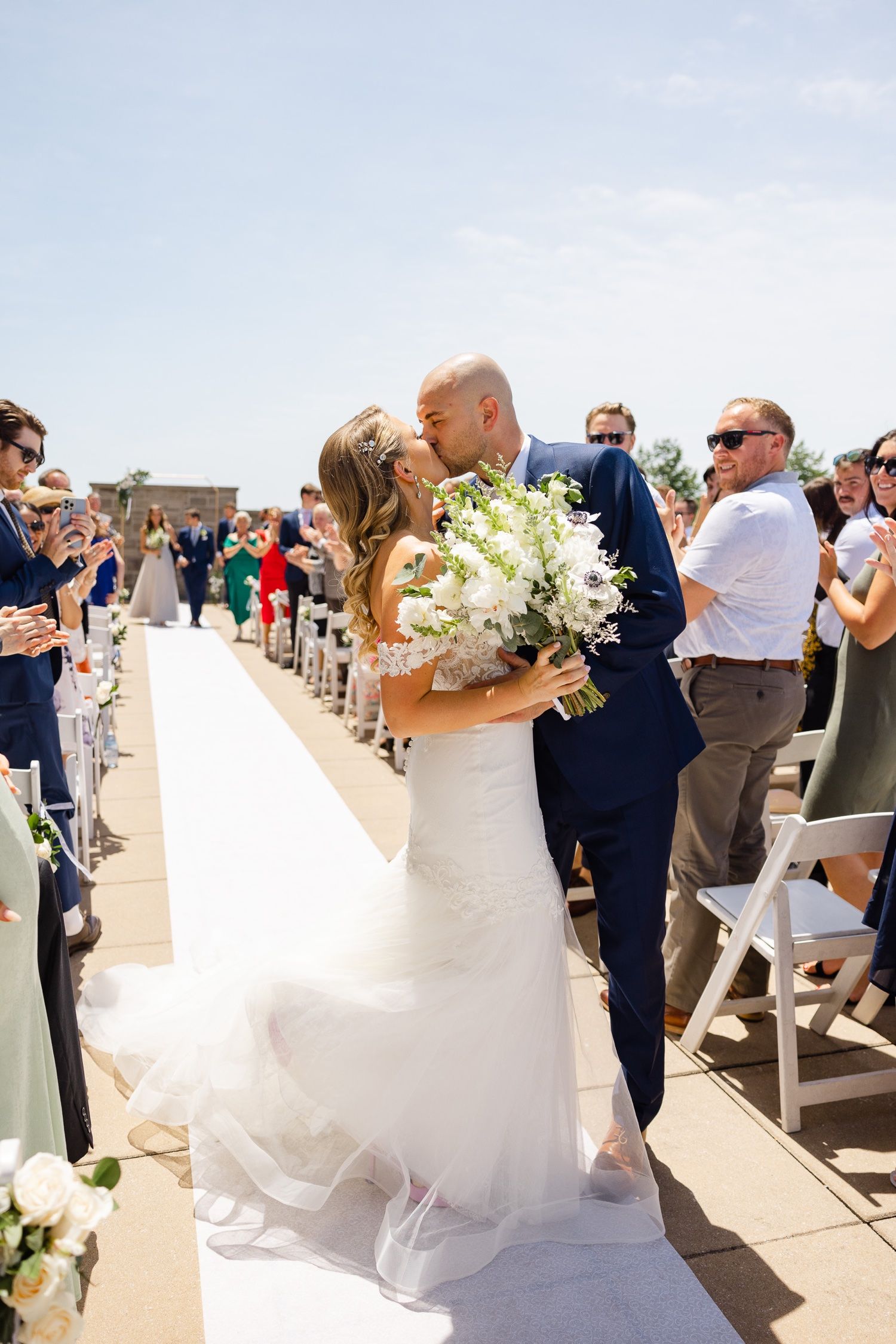 Wedding celebration on sunny rooftop terrace with guests looking on as couple shares first kiss.