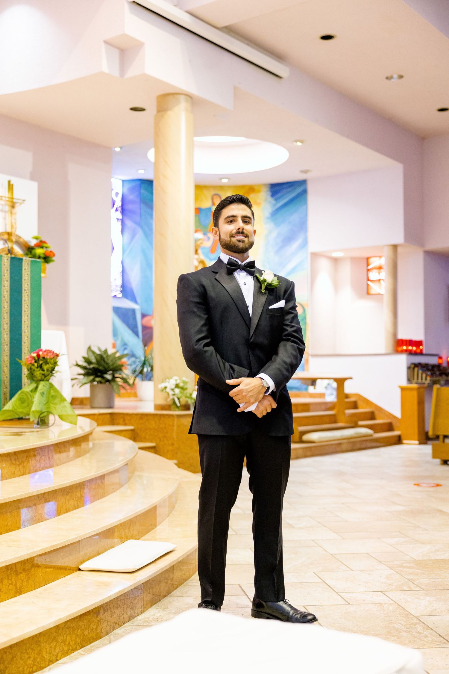 Groom in black tuxedo poses at altar with decorated church sanctuary in background.