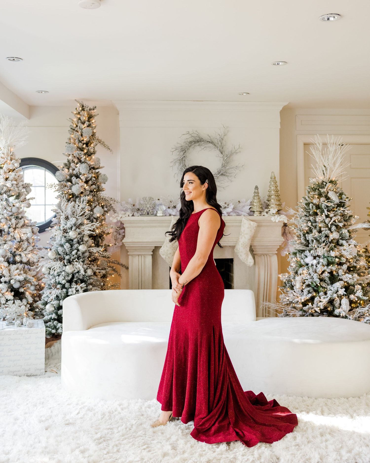 Woman in flowing red gown poses in white room with beautifully decorated Christmas trees.