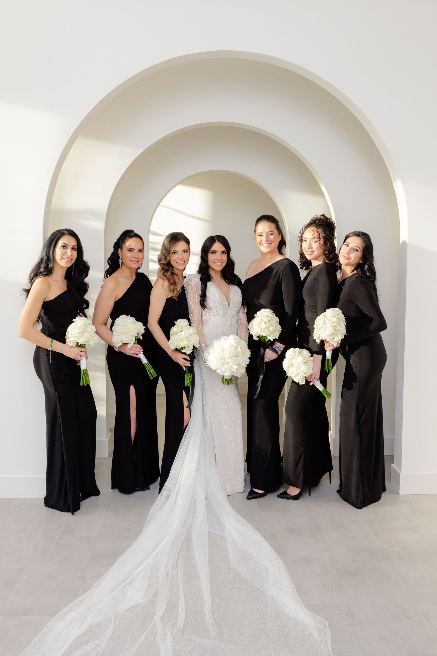 Wedding party with bridesmaids in black dresses holding white bouquets under architectural arches.