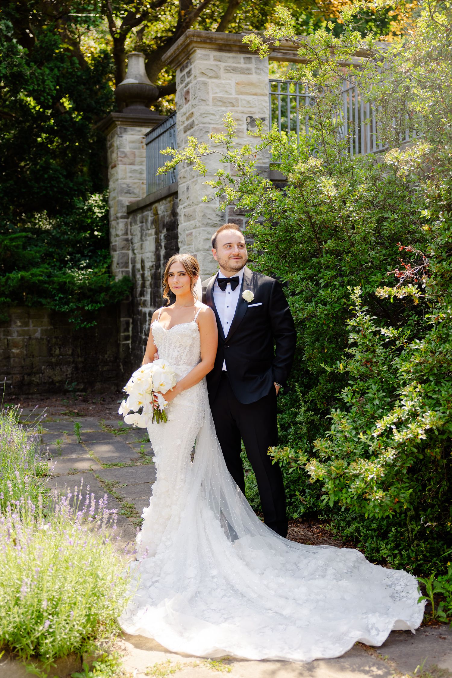 A couple in formal wedding attire poses together in a garden setting with stone architecture and blooming flowers.