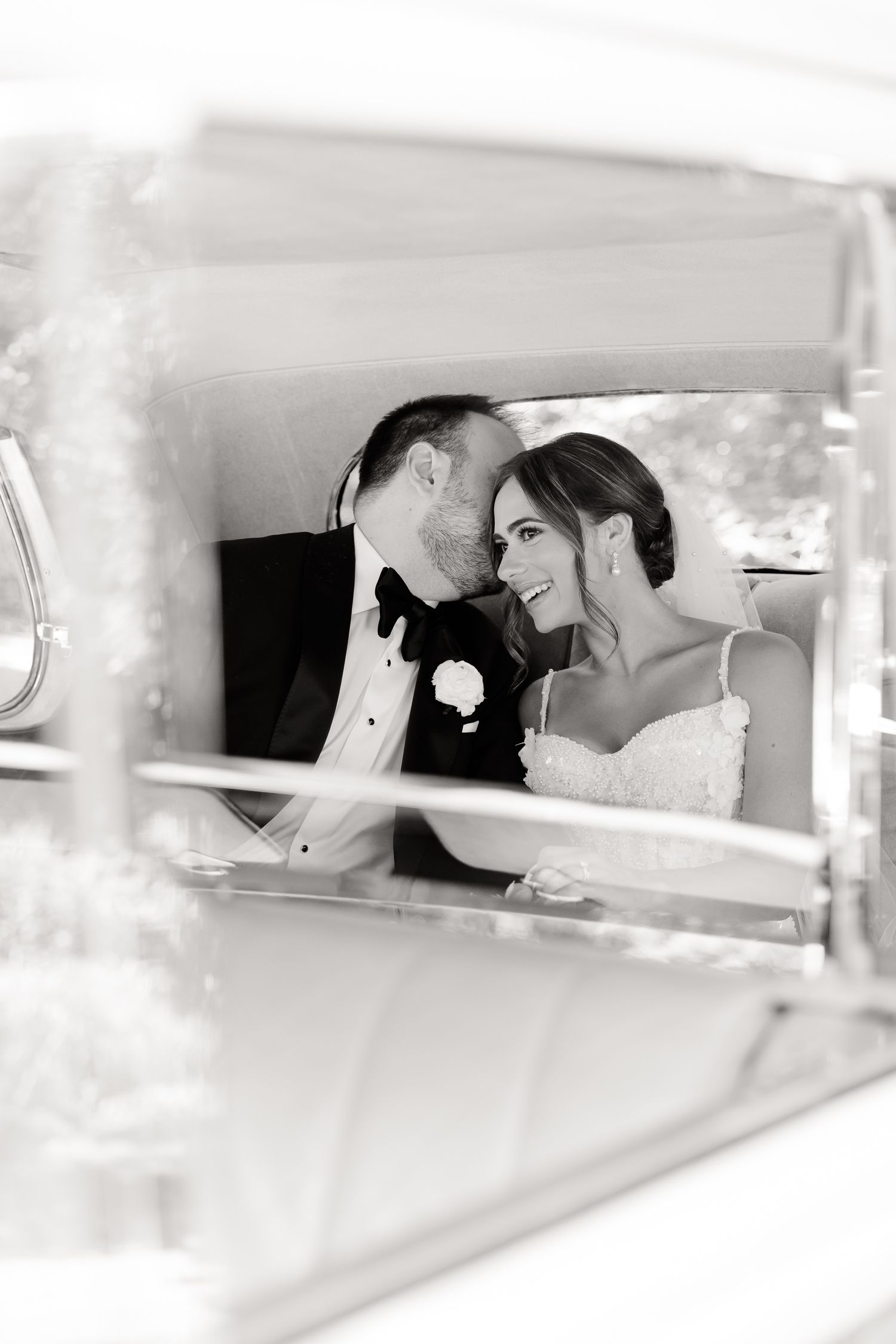 A romantic black and white photo of newlyweds sharing an intimate moment in the back seat of a vintage car.