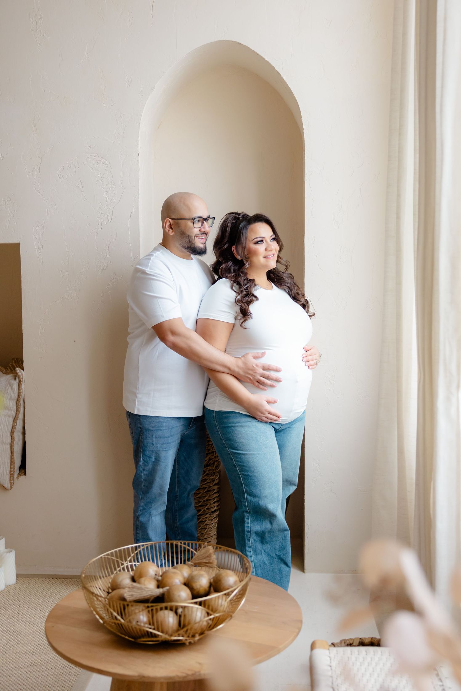 A tender maternity moment showing a couple in white tops and jeans standing in an arched doorway.