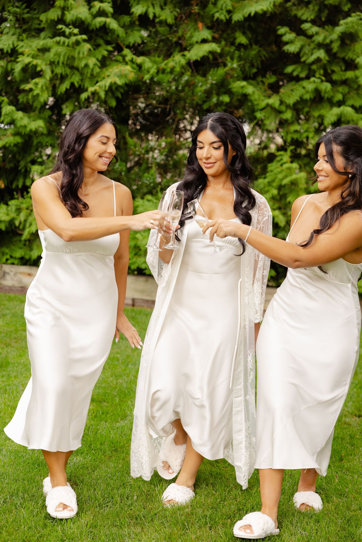 Three women in flowing white dresses sit together on grass, enjoying a casual outdoor moment.
