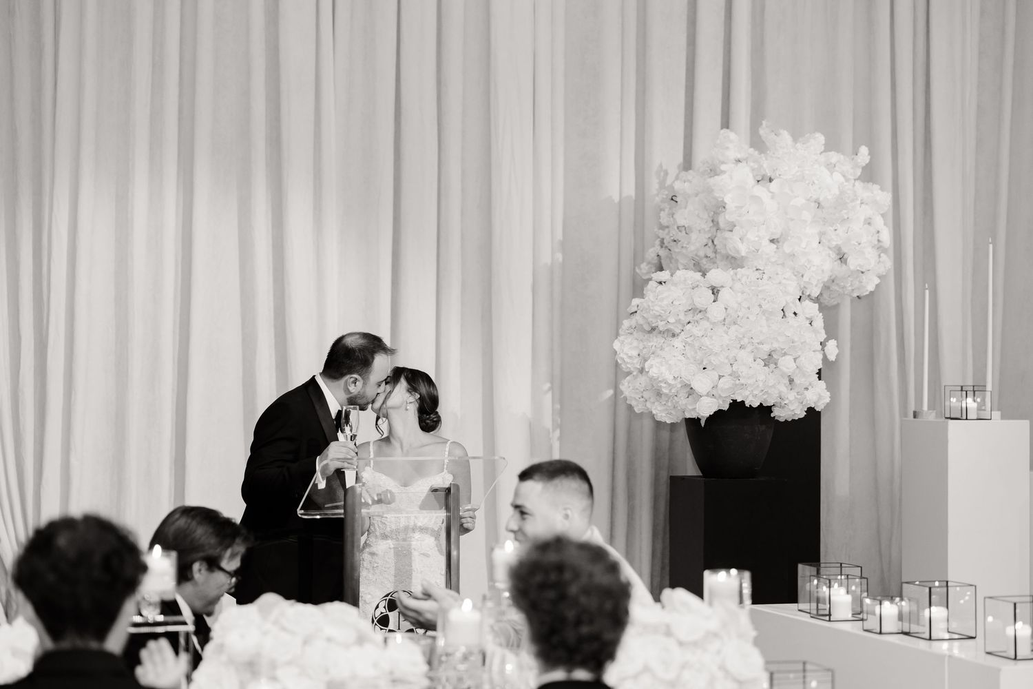 Black and white panoramic shot of an elegant wedding reception with white floral centerpieces and draped curtains.