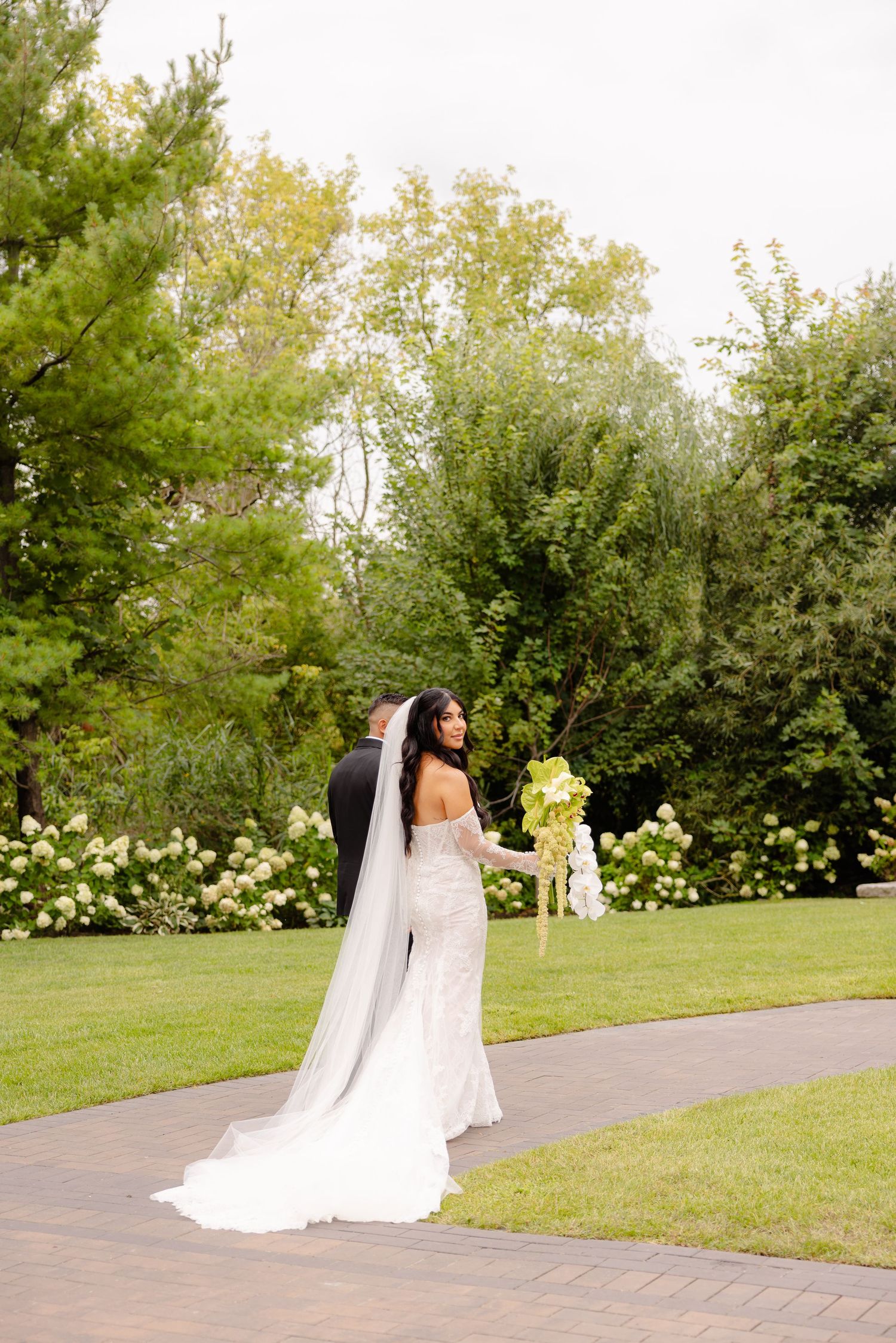 A bride in a flowing white wedding gown and veil walks along a garden pathway on her special day.