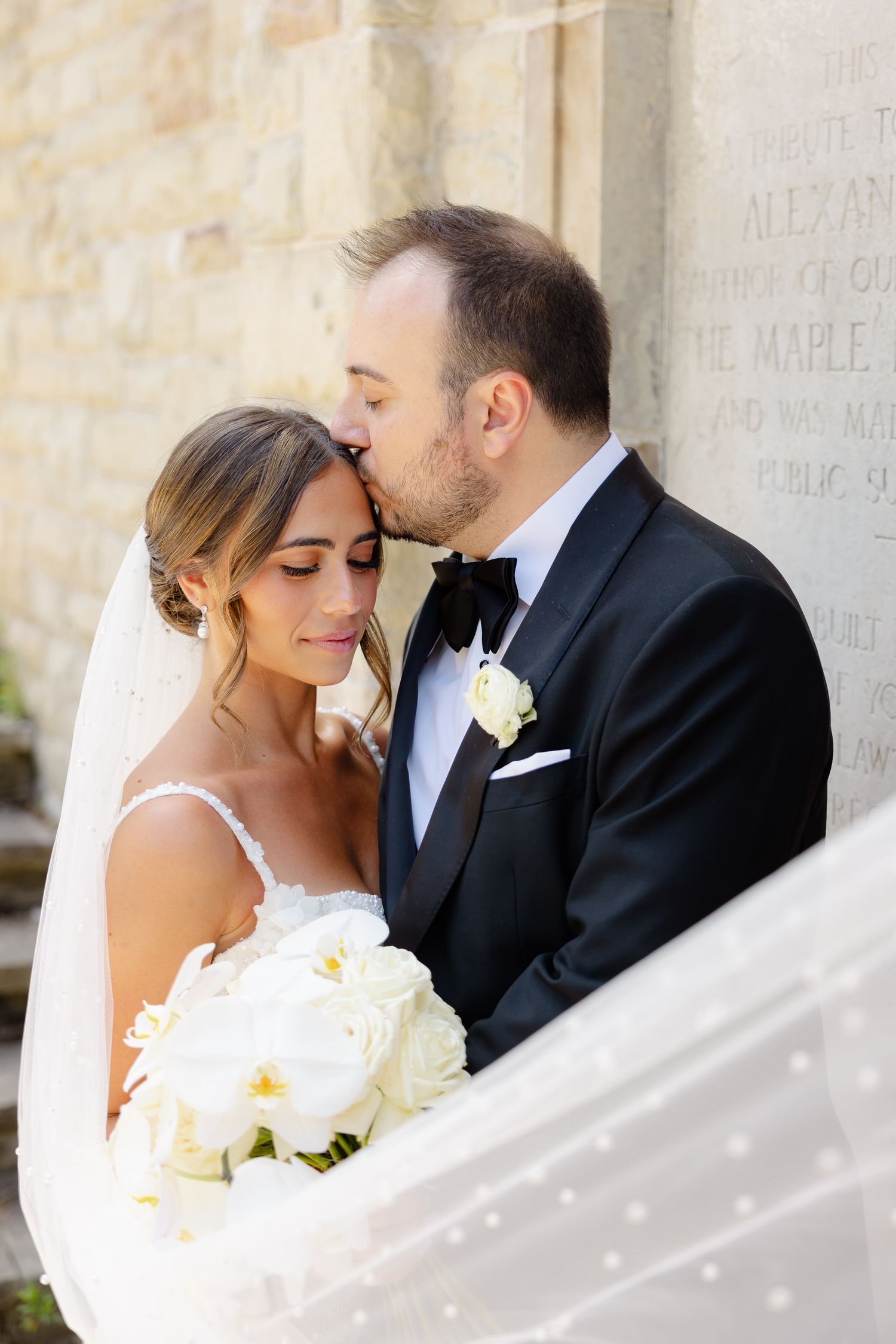 A bride and groom share an intimate moment against a white stone wall backdrop, with the bride holding white roses.