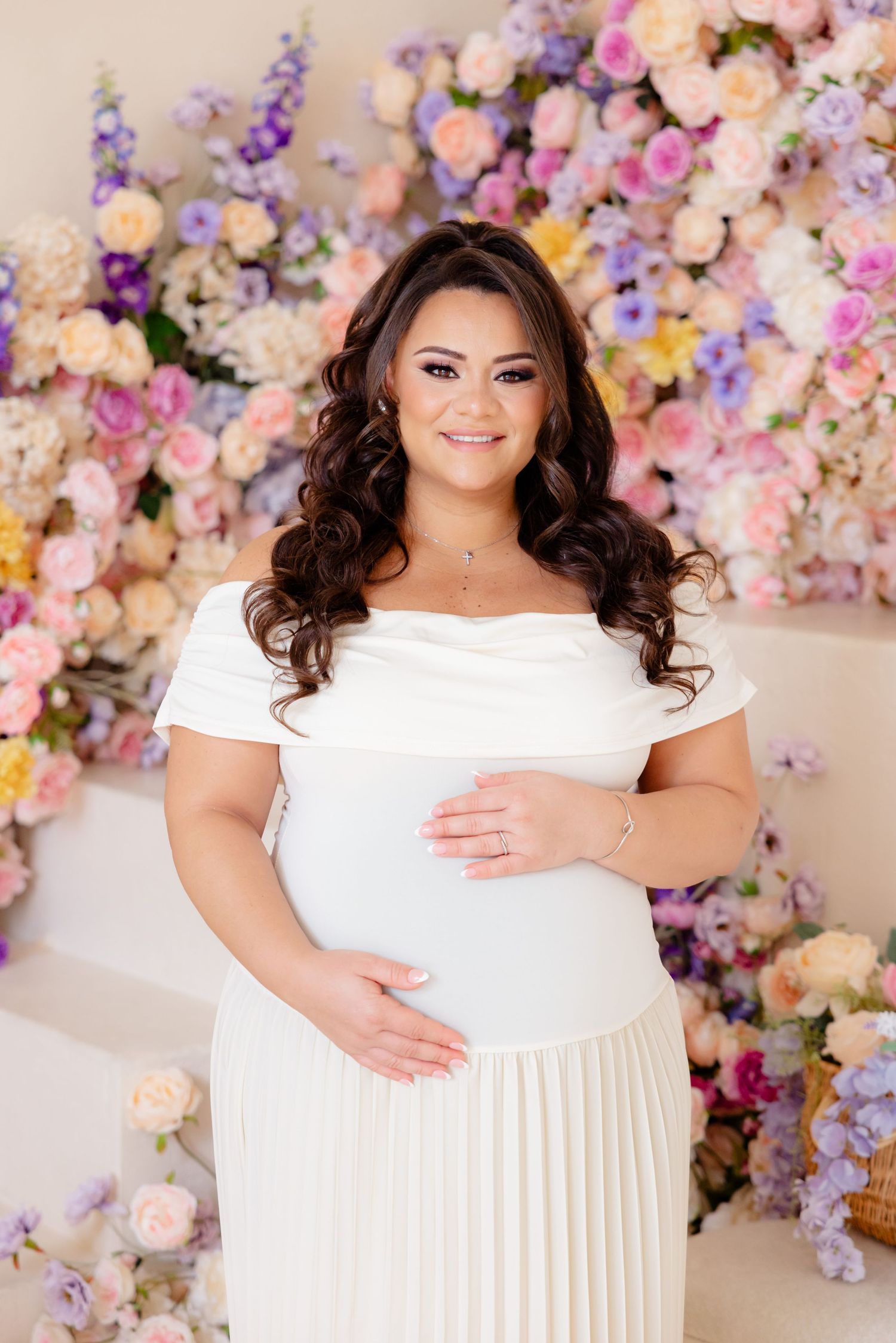Maternity photo with beautiful floral backdrop featuring pink and purple blooms behind a person in an elegant white dress.