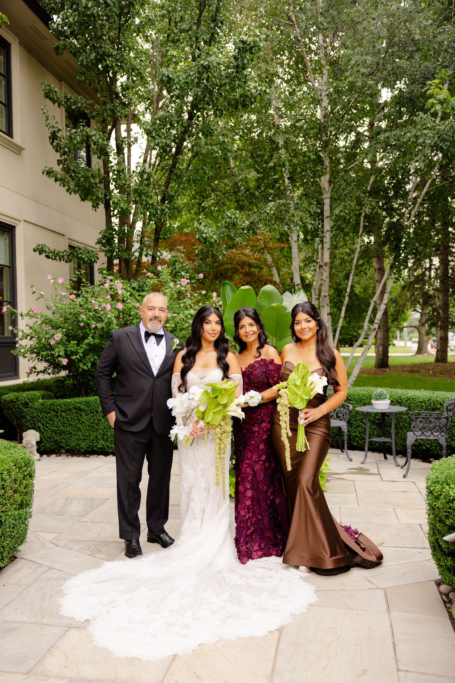 A wedding party poses outdoors on a stone patio surrounded by lush green trees and gardens.