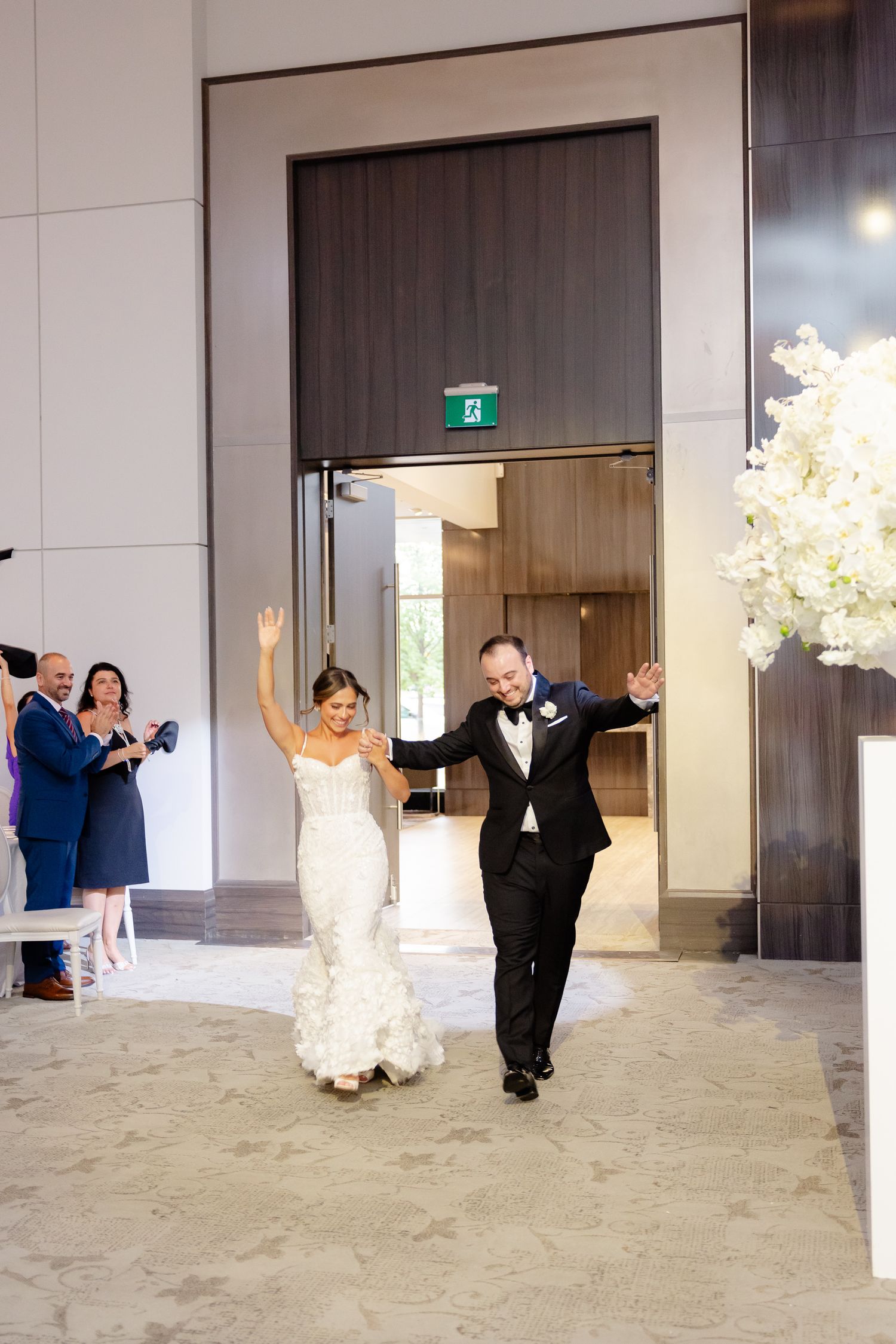 Newlyweds make their grand entrance at a modern venue, raising their arms in celebration while guests cheer.
