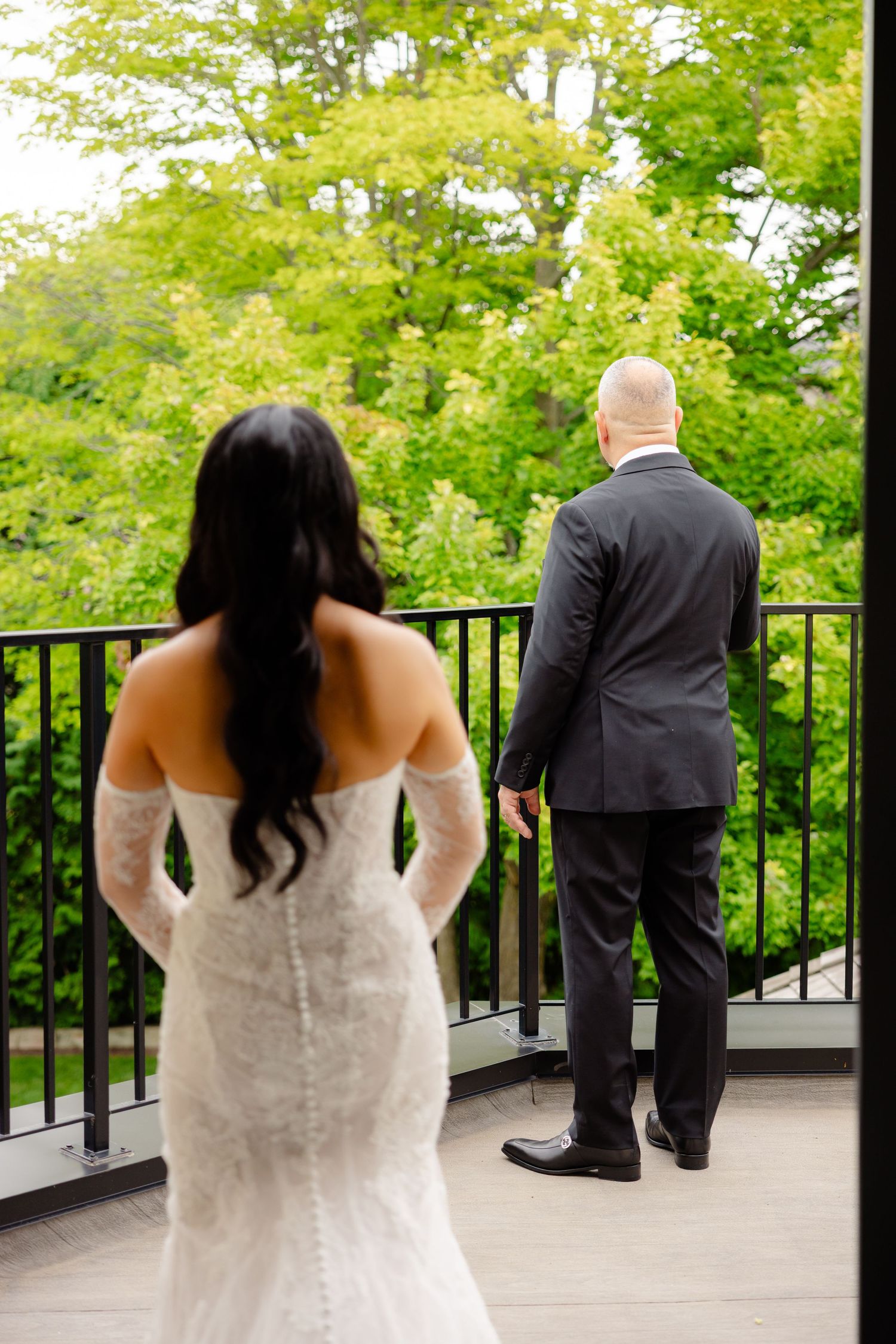 A wedding couple shares a traditional first look moment on an outdoor balcony surrounded by lush green trees.