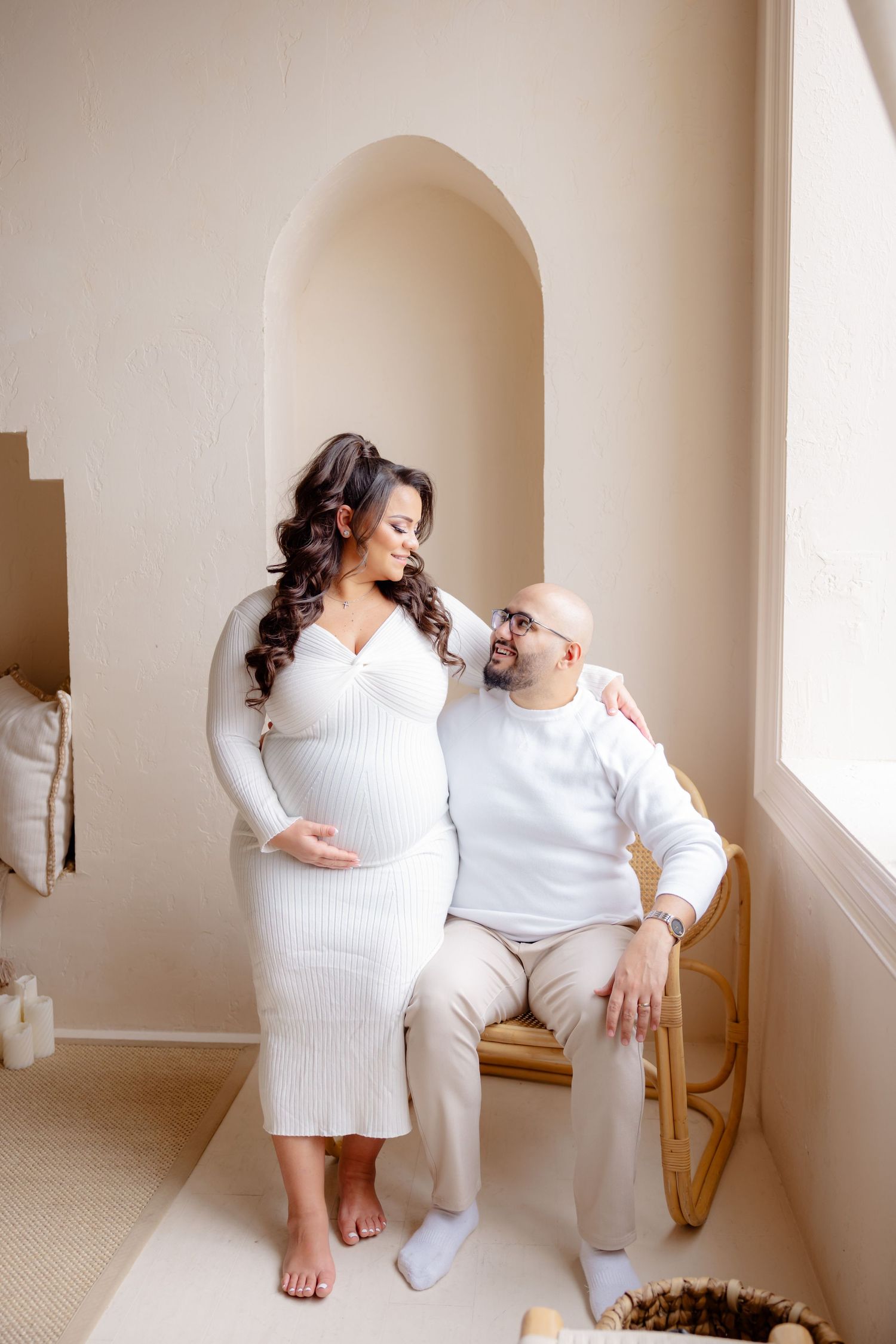 A couple wearing all white outfits sits together by a window in a minimalist room with arched details.