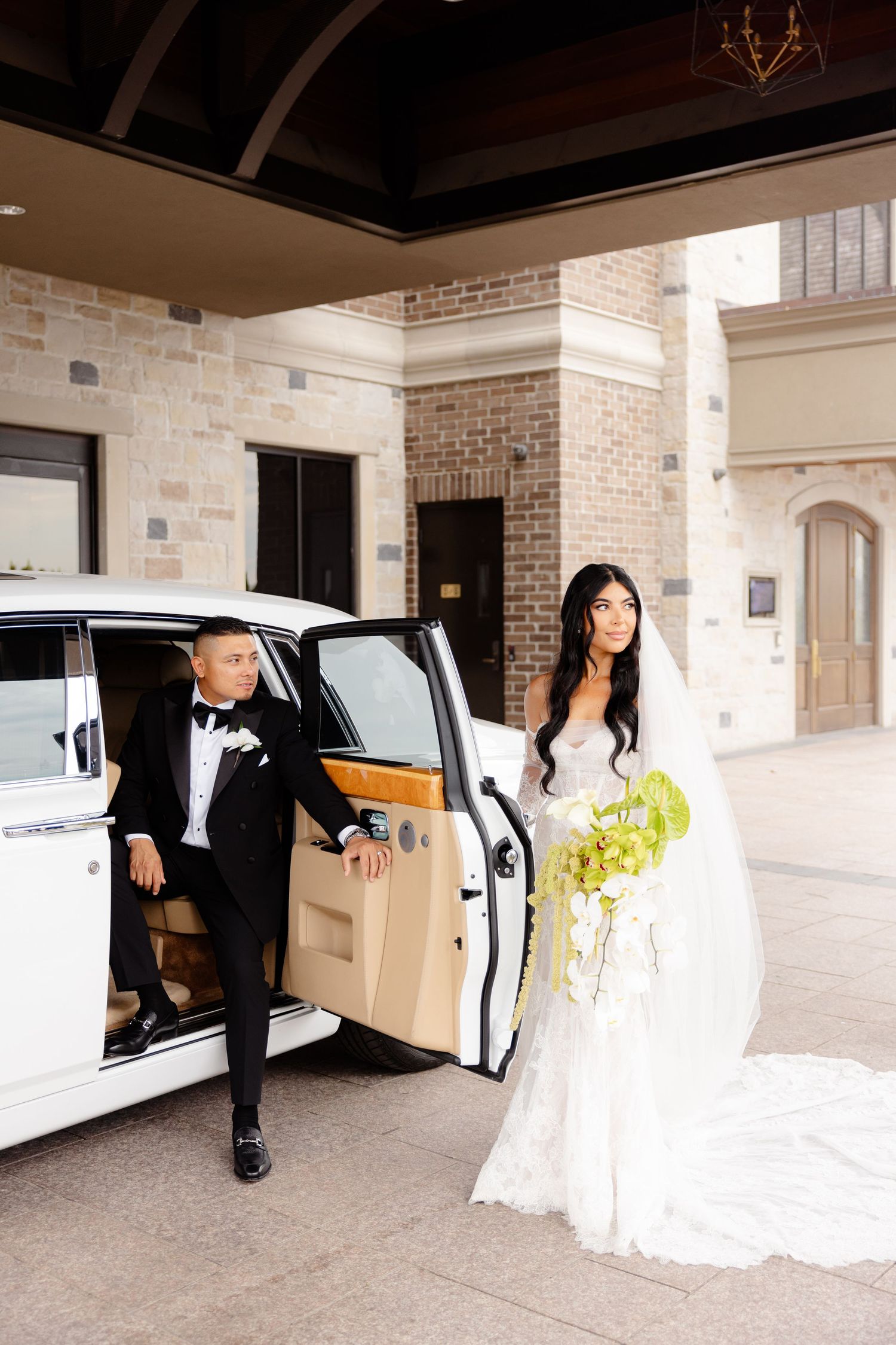 Wedding party entering and exiting a white luxury car in front of a brick facade venue during a formal celebration.