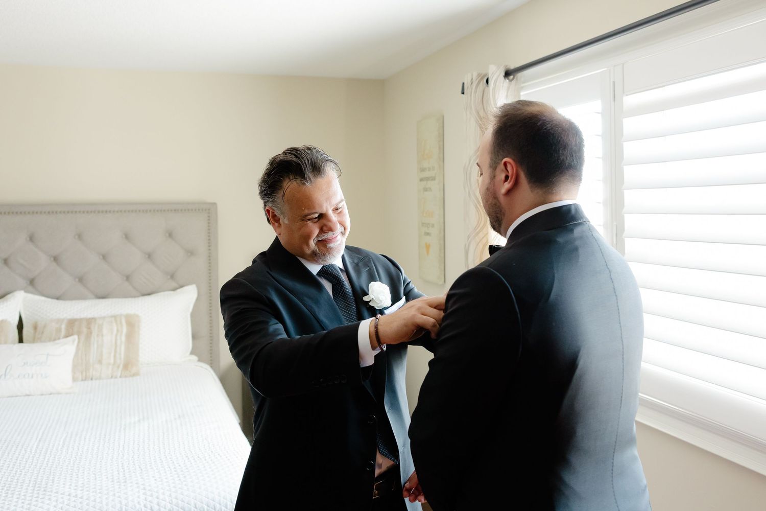 Two people in black suits help each other with formal attire adjustments in a bright bedroom setting.