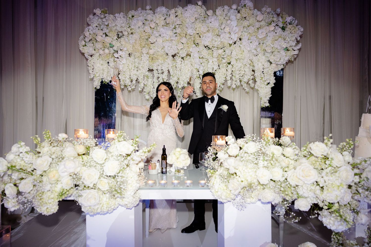 Wedding couple stands at decorated table with stunning white floral backdrop and arrangements.