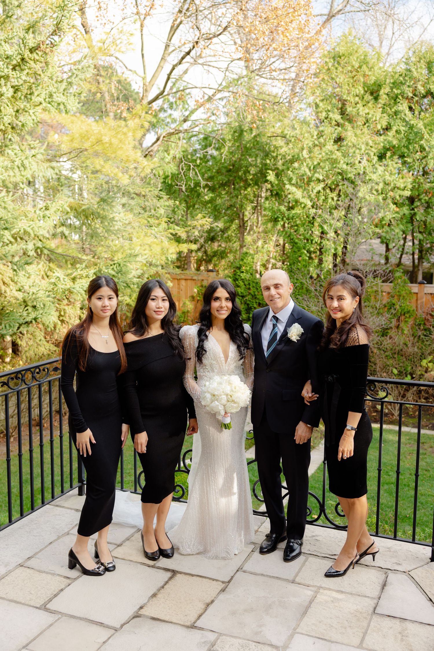Wedding party in black attire poses with couple on stone terrace with garden setting.