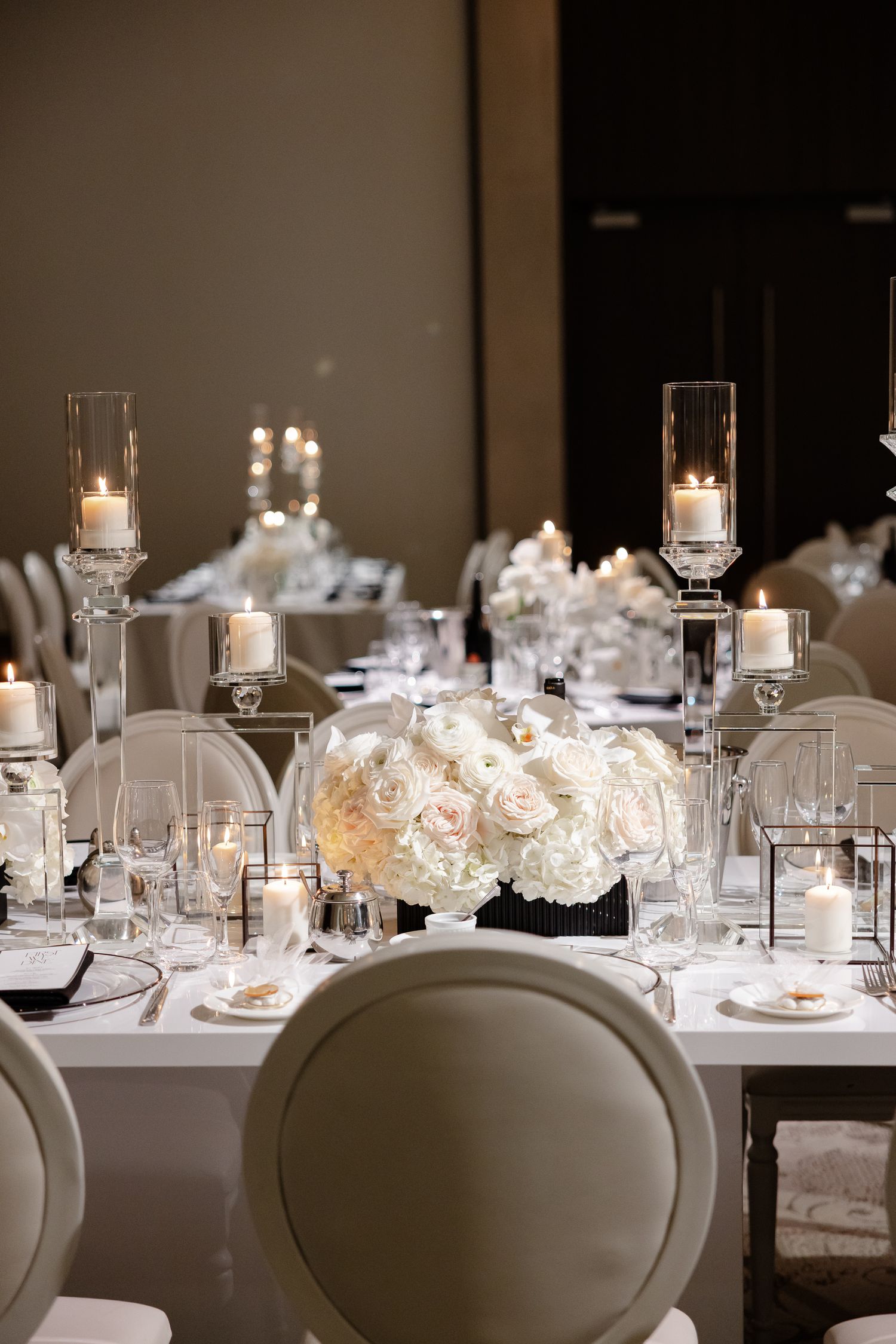 Elegant wedding table setting with white roses, glass candleholders, and modern place settings in a formal dining room.